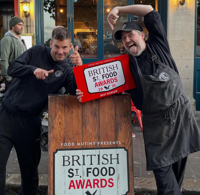 Two men posing with a sign at the British Street Food Awards, one giving a thumbs up and the other making a playful gesture. They are outdoors in front of a building with large windows, with other people in the background.