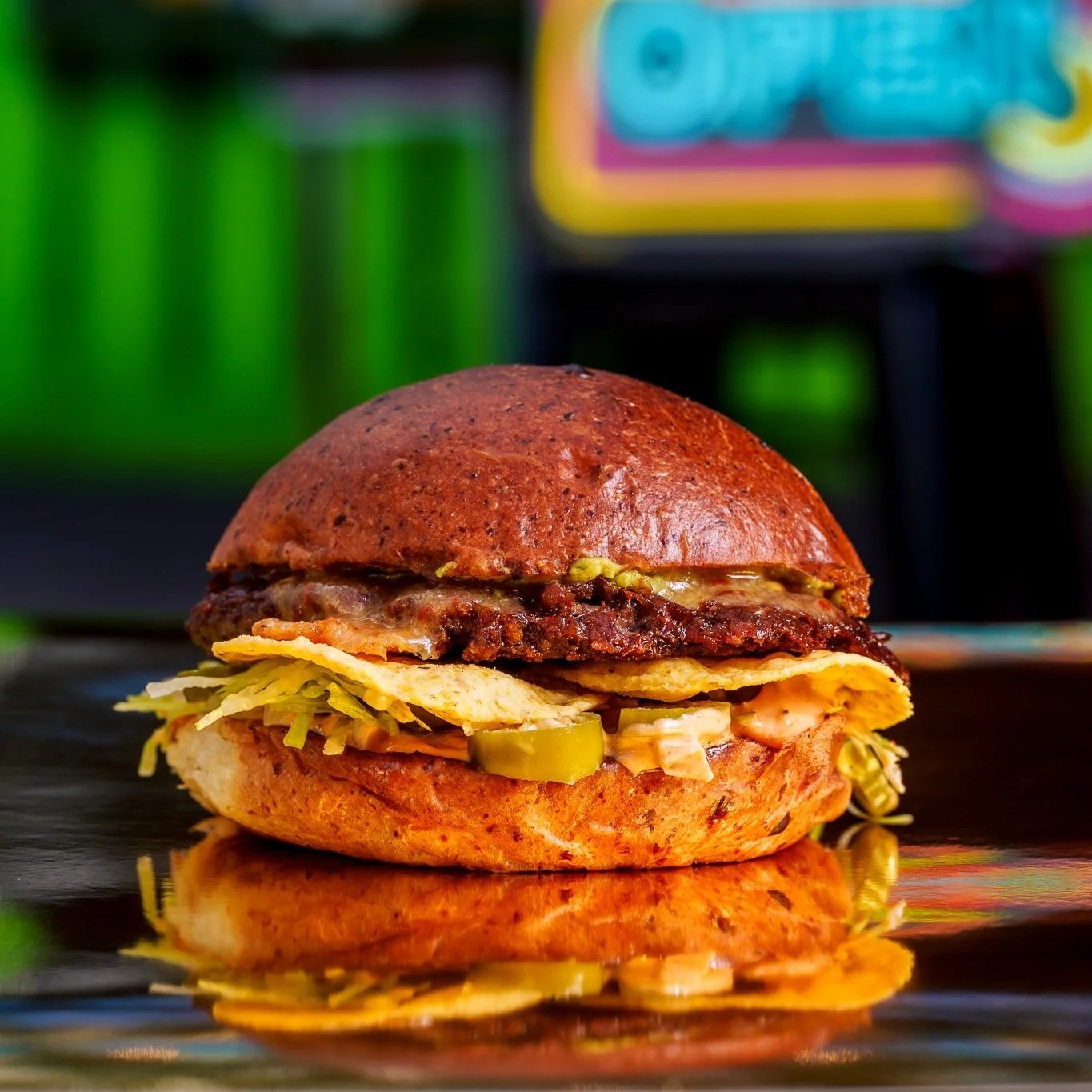 A burger with a toasted bun, lettuce, pickles, a slice of tomato, melted cheese, a beef patty, bacon, and a fried egg, with a colorful background.