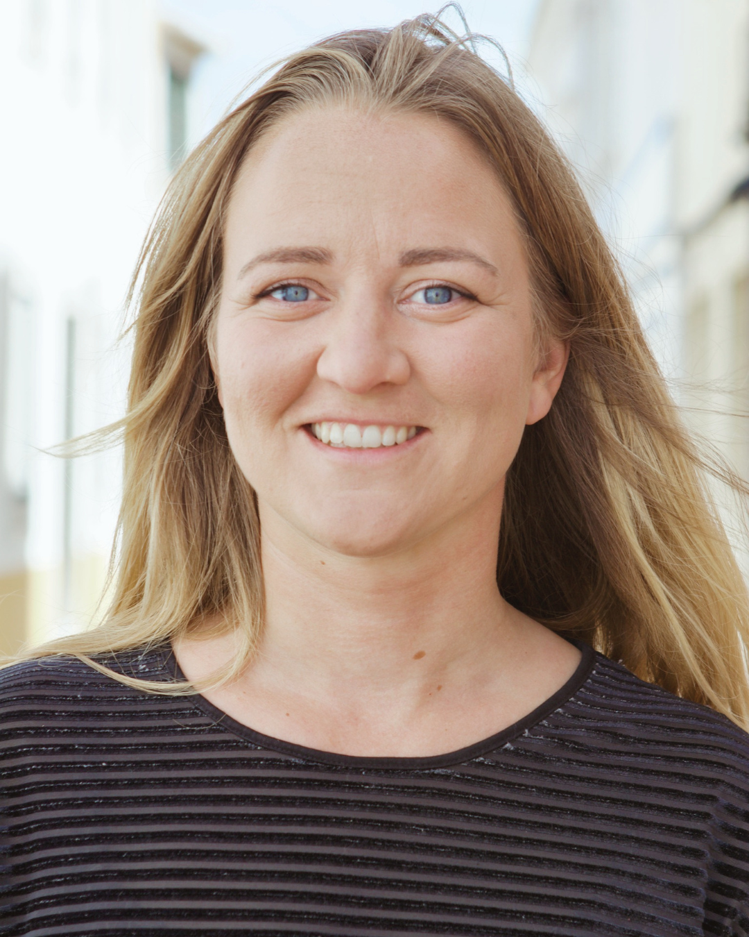 Close-up of smiling woman with blue eyes and blonde hair wearing a black striped shirt in a bright indoor setting.