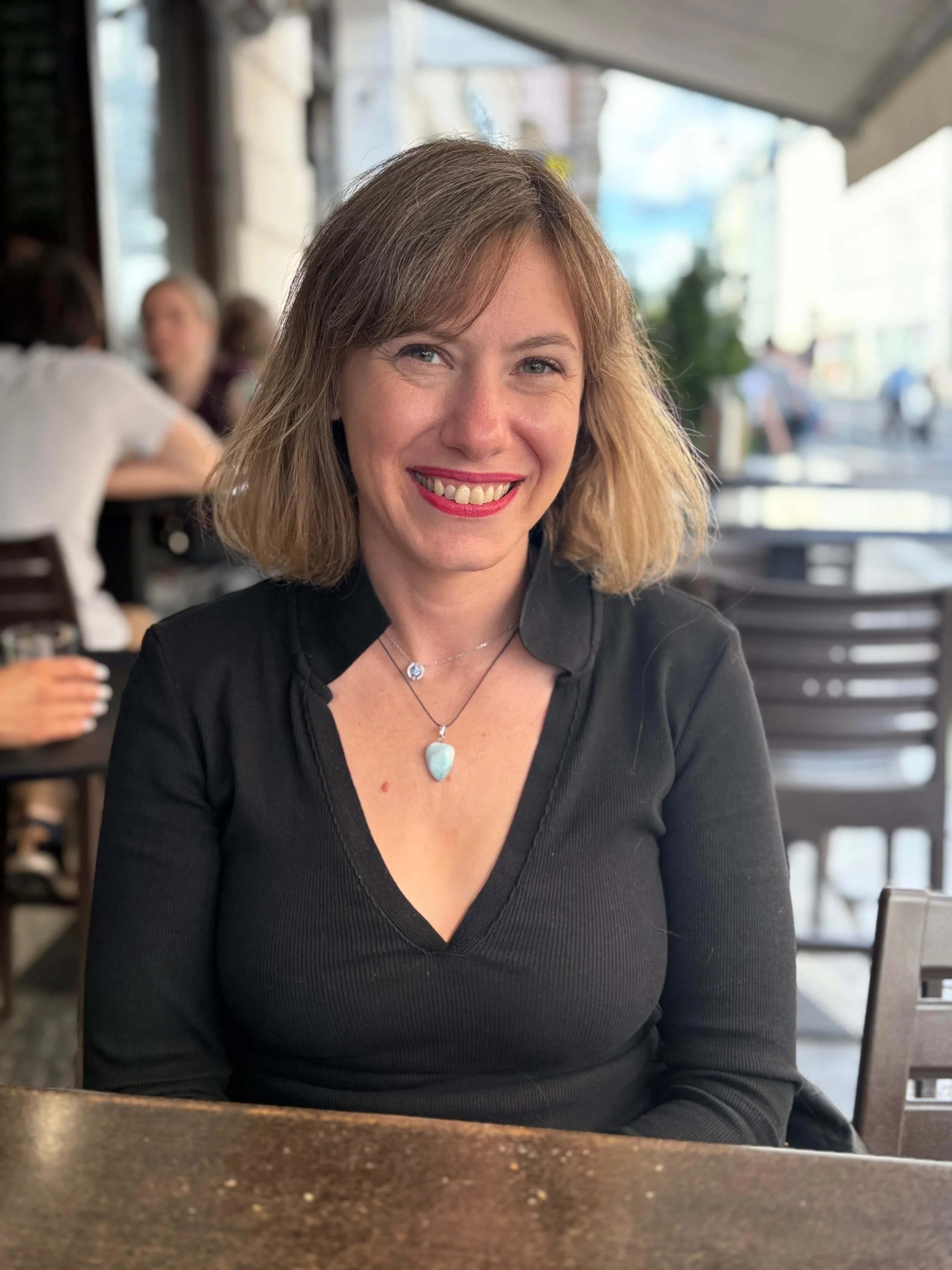A woman with shoulder-length blonde hair, wearing a black top and jewelry, is smiling and sitting at a restaurant or cafe with a blurred background of other people and city streets.