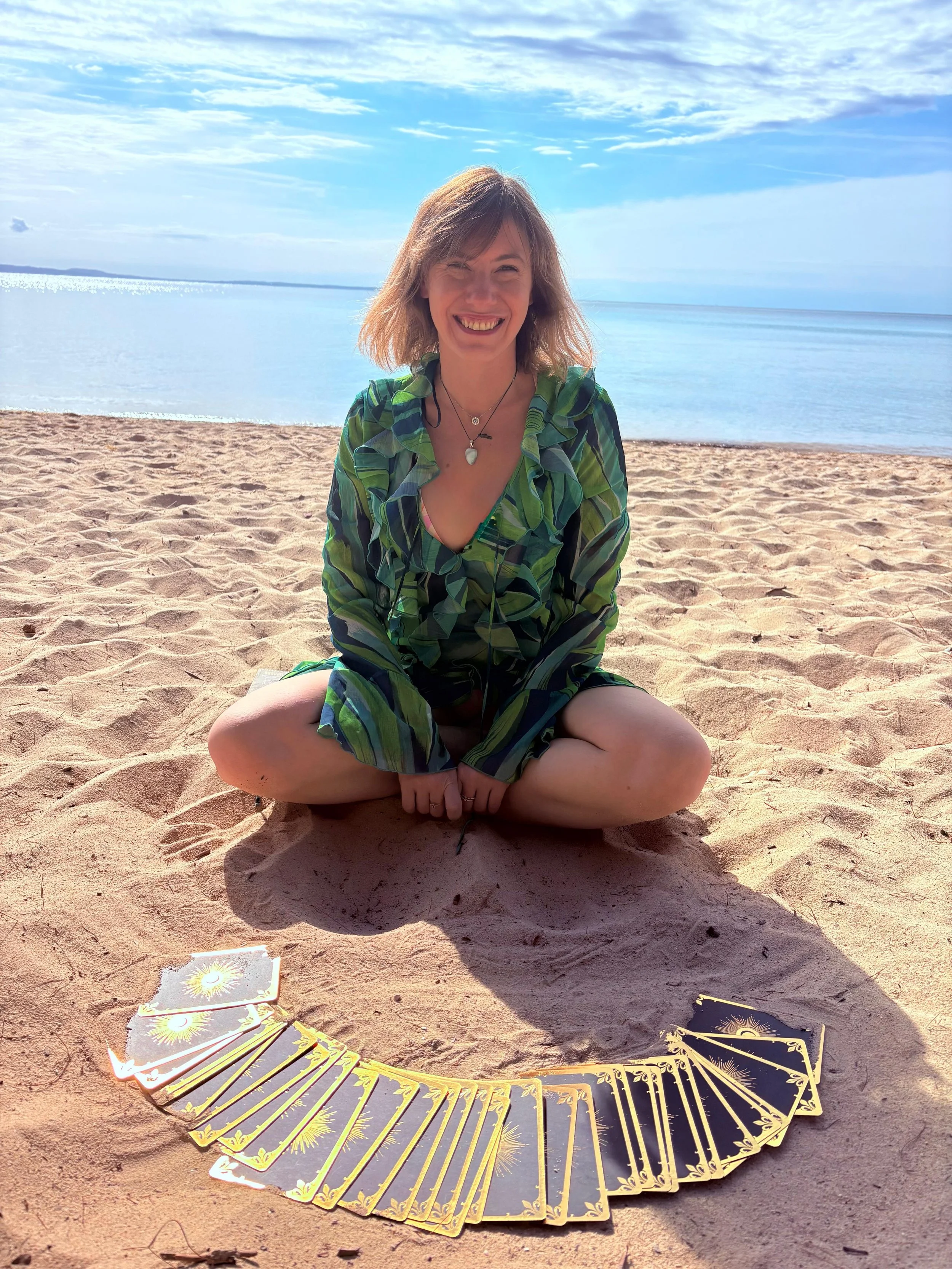 A woman sitting cross-legged on a sandy beach, smiling, with Tarot cards laid out in a semi-circle in front of her, overlooking calm water and a partly cloudy sky.