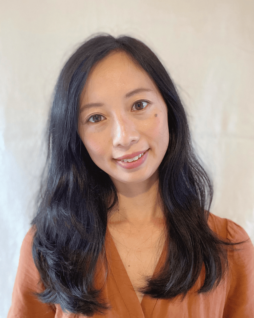 A woman with long, wavy black hair smiling against a light-colored background.