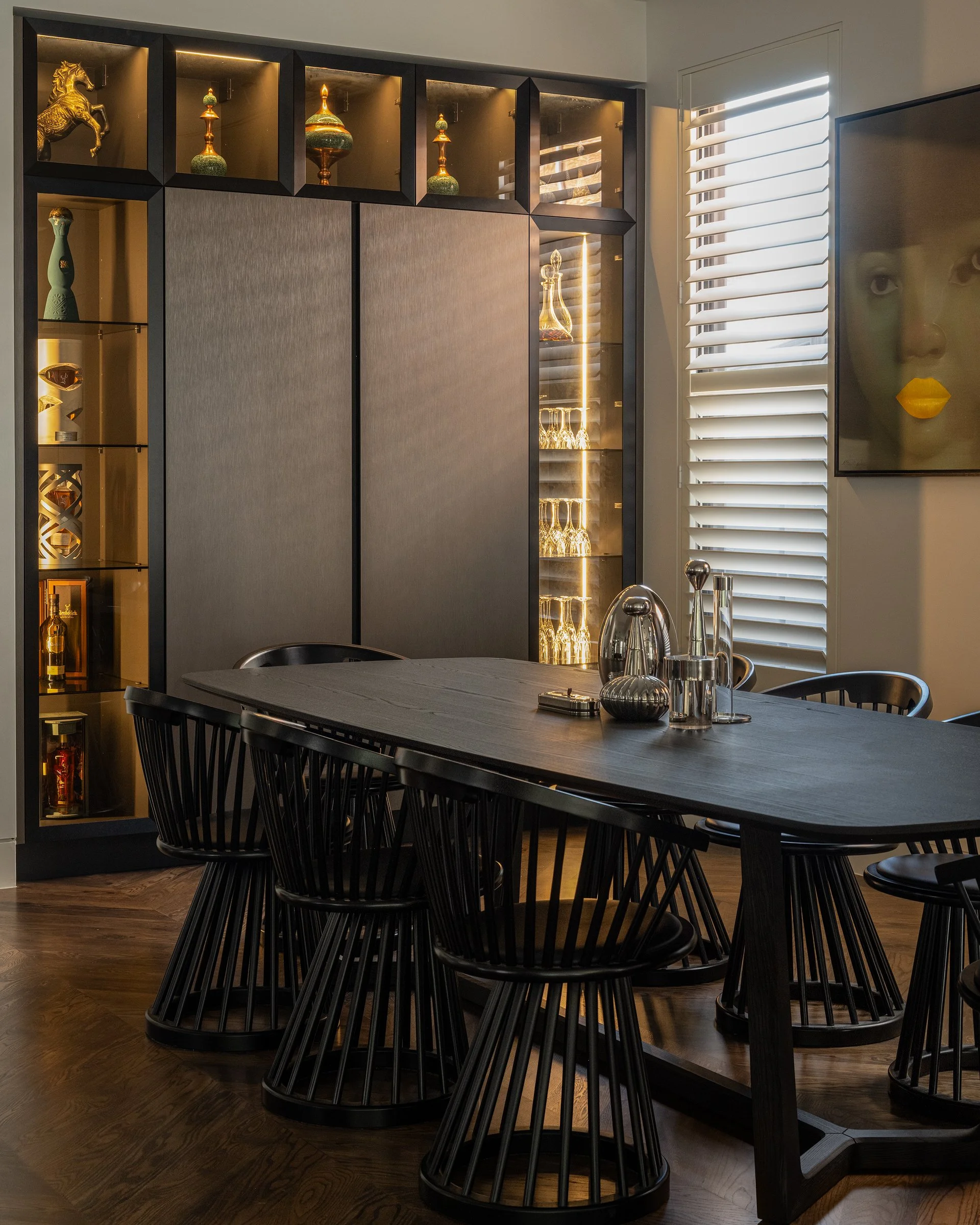 Modern dining room with a dark wooden table, black spindle-back chairs, a glass cabinet with decorative items and glassware, a window with white blinds, and a framed artwork of a face with yellow lips on the wall.