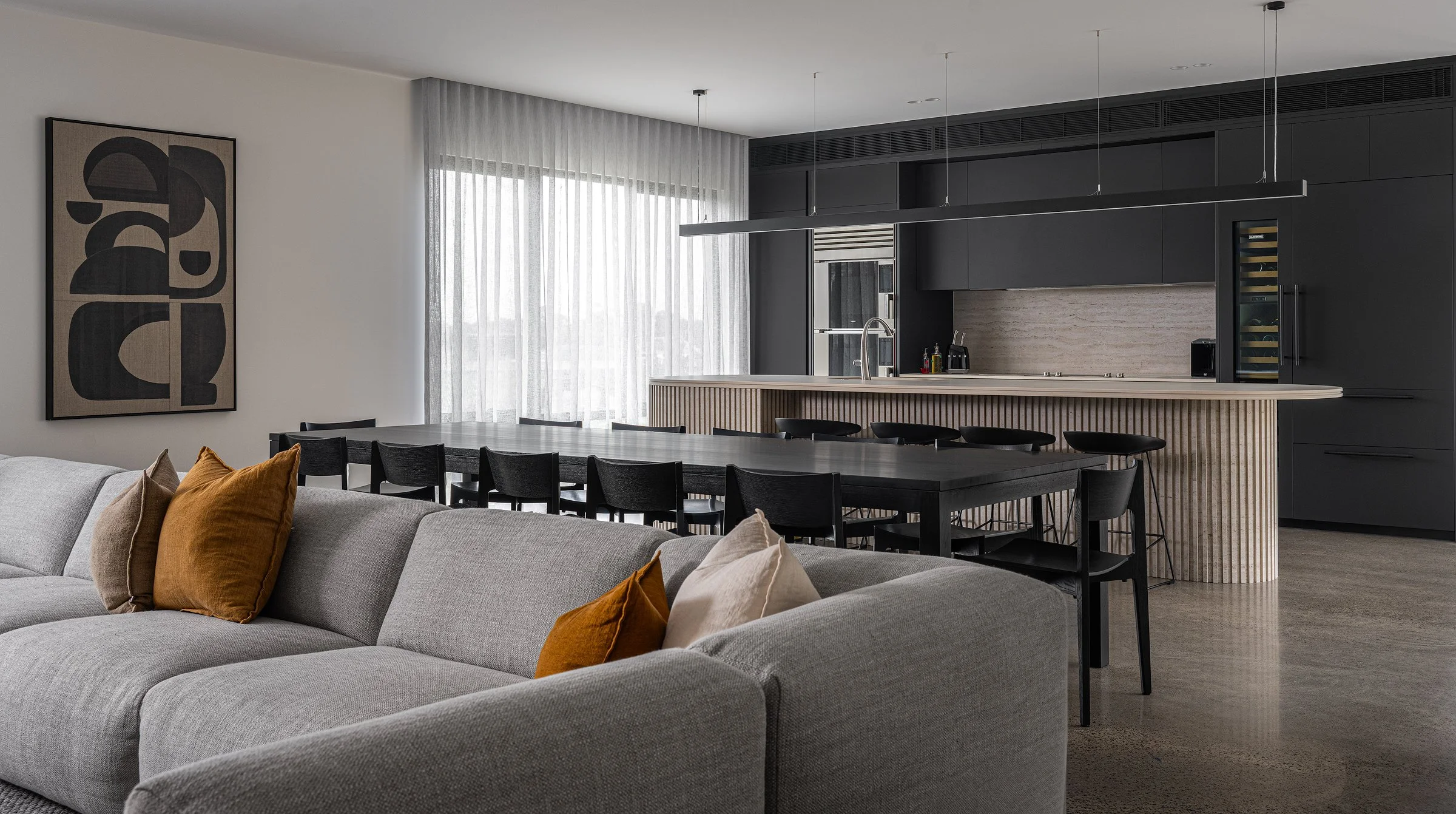 Modern open-plan living room and kitchen with gray sofa, orange and beige pillows, black dining table with chairs, black kitchen cabinets, light wood kitchen island, large window with sheer curtains, abstract wall art.