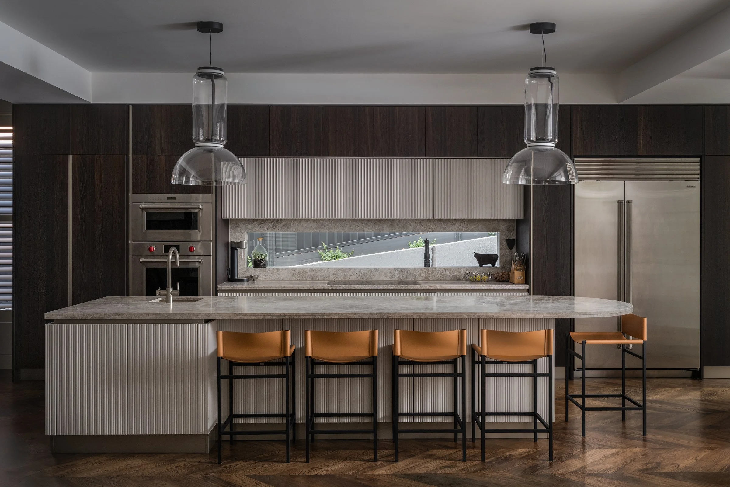 Modern kitchen with dark wood cabinets, a marble island with beige barstools, and stainless steel appliances, including a refrigerator and wall oven, with a long horizontal window above the counter.