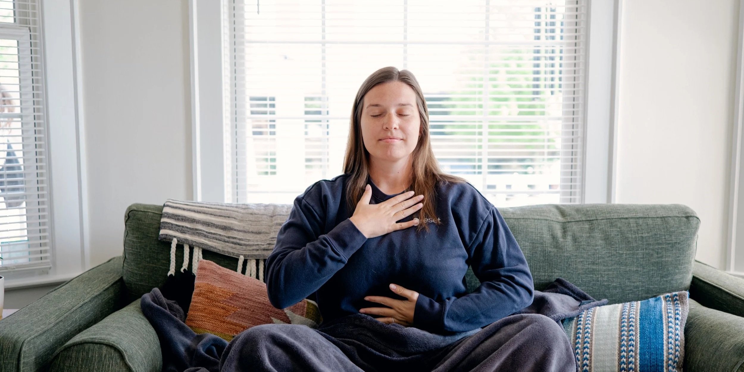 Woman sitting on a green sofa with her eyes closed, hand on her chest, and another on her stomach, practicing meditation or mindfulness in a bright living room.