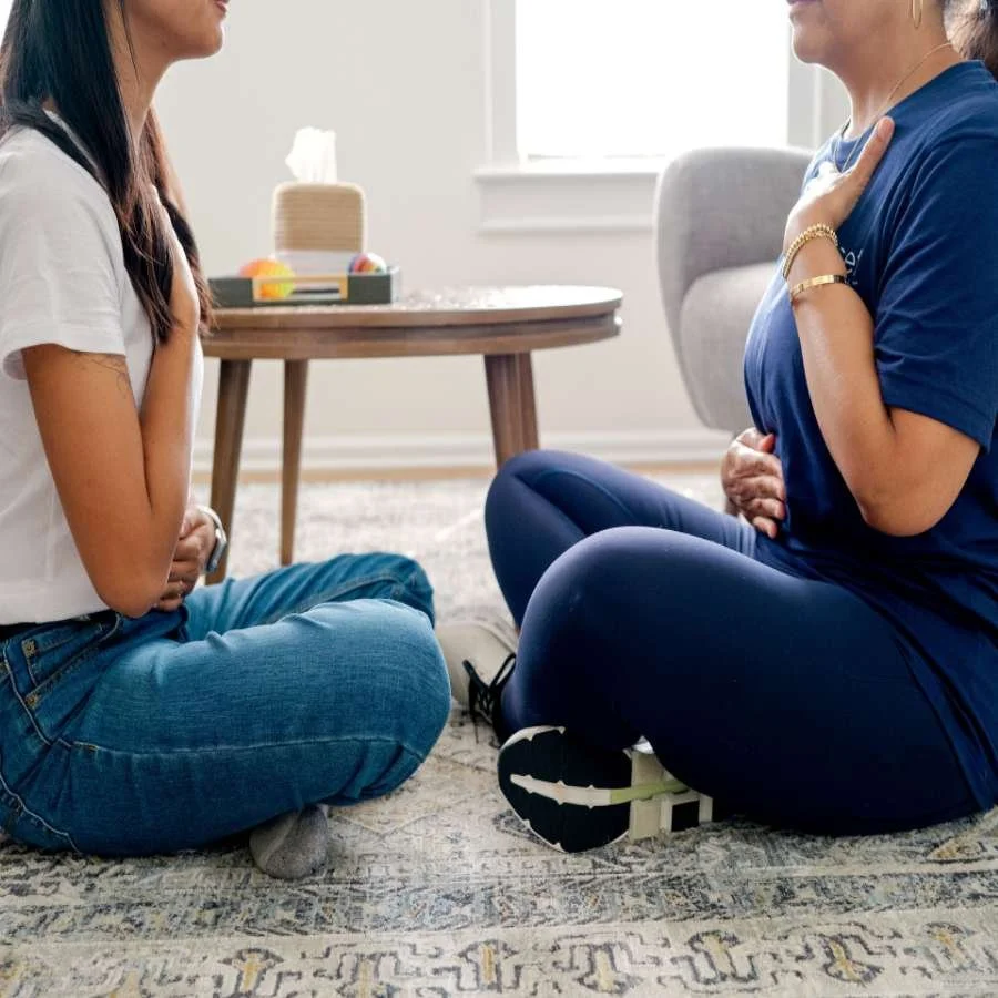 Two people from different racial backgrounds sit on the ground, facing each other, and participate in a grounding and coregulating exercise.