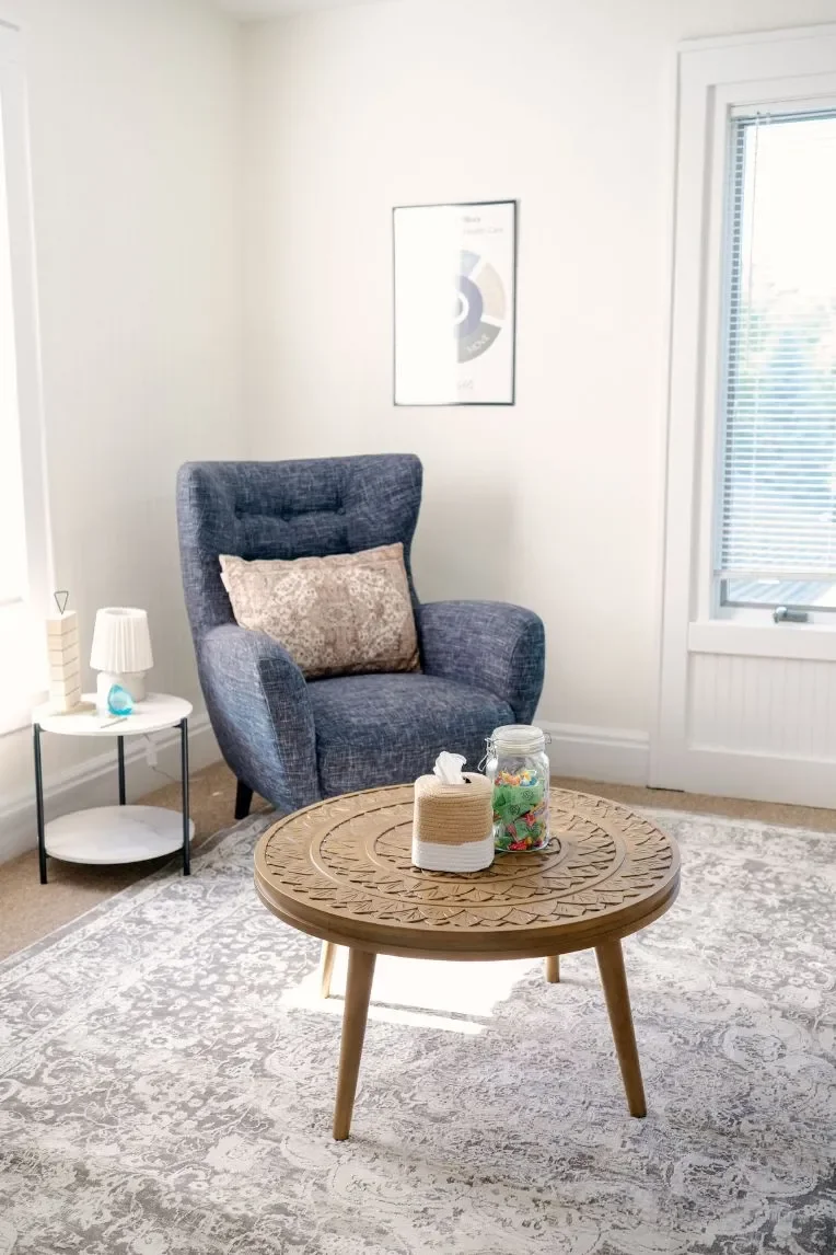 A cozy Reset office with a gray armchair, decorative pillow, small side table with lamp and decorative items, round wooden coffee table with tissue box and jar of colorful candies, patterned area rug, and a window with blinds.