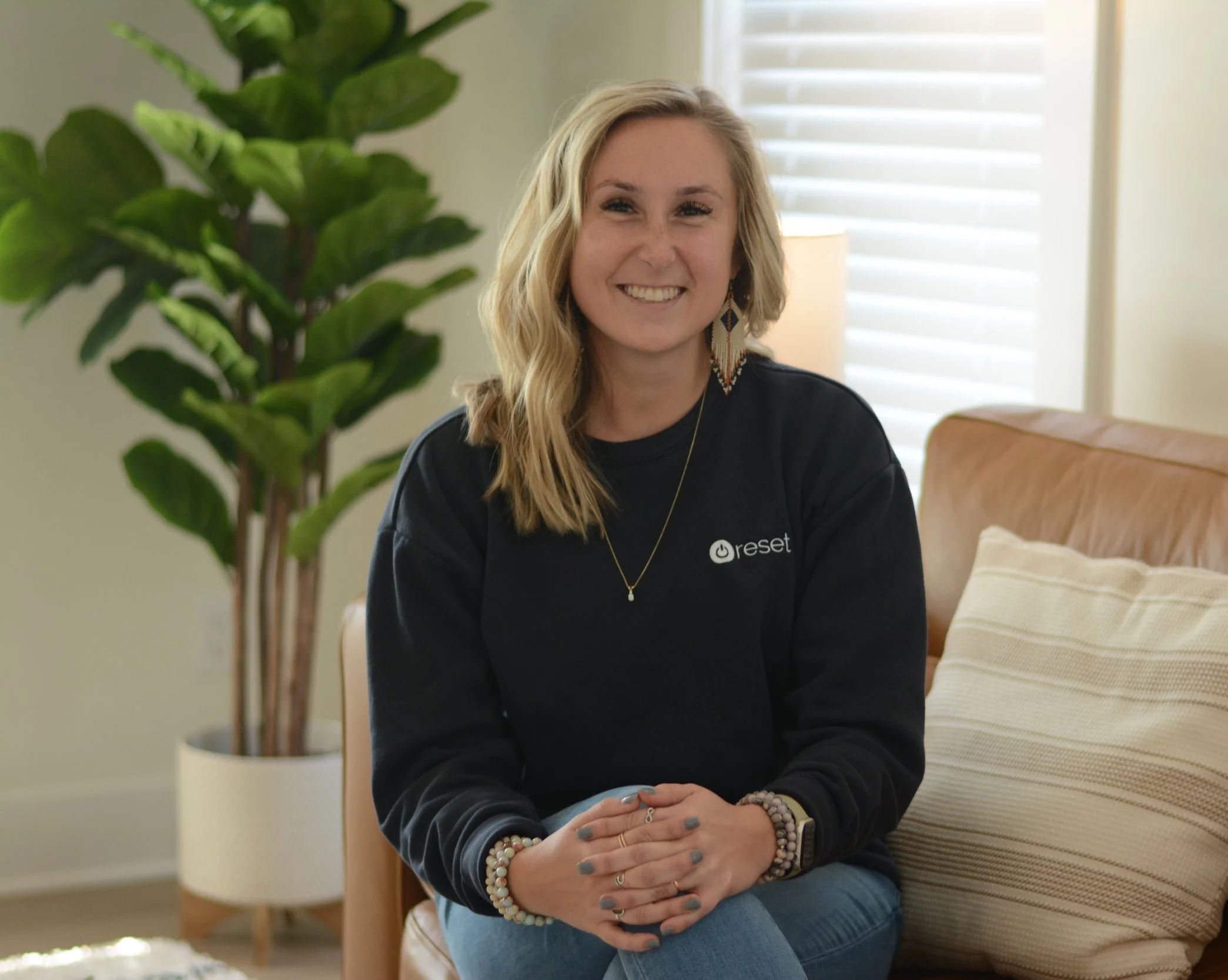 A young woman with blonde hair smiling while sitting on a couch in a cozy living room, wearing a sweatshirt with a logo that says 'reset', accessorized with earrings, bracelets, and a necklace, with a large green plant and window with blinds in the background.