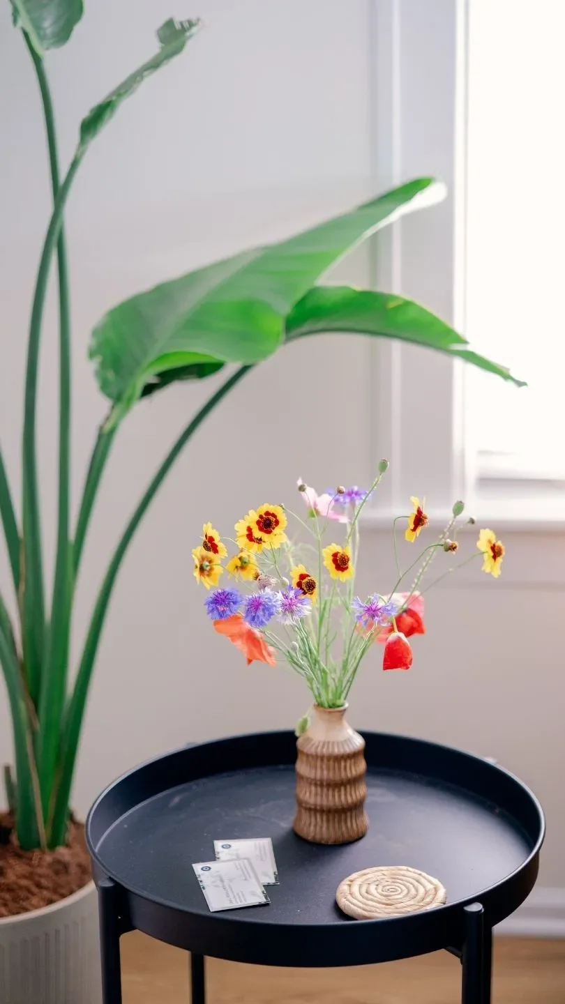 In a Reset office, there is a black round side table with a small floral arrangement. Behind the table is a large green potted plant, and in the background, there's a window with natural sunlight.
