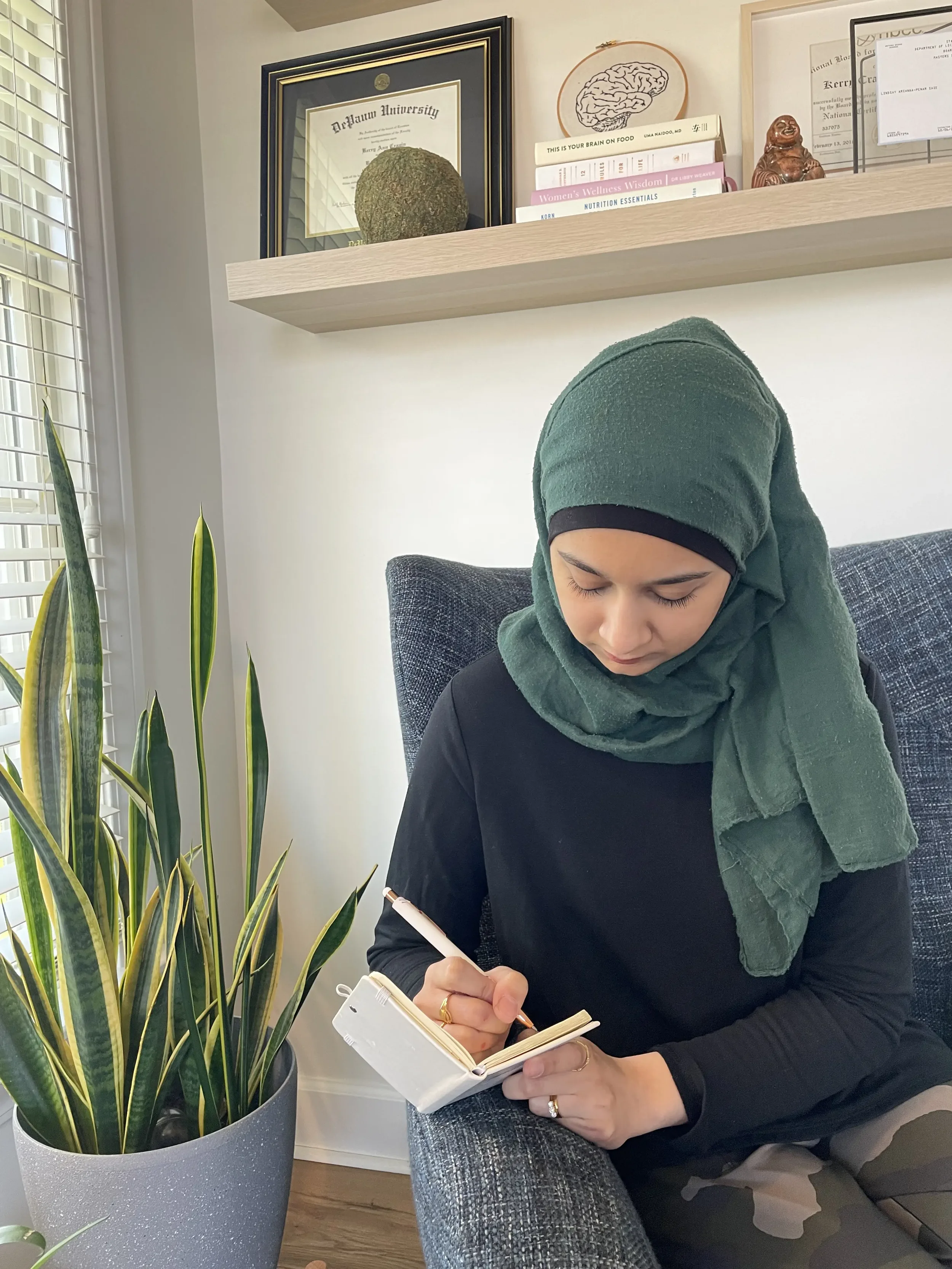 A woman wearing a hijab writes in a journal. She is seated in a dark blue chair next to a potted plant.