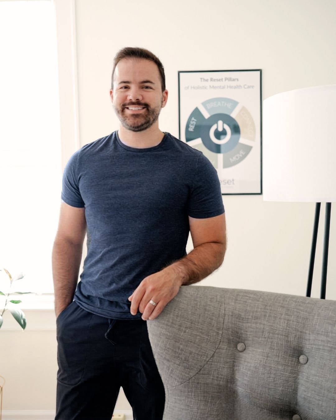 Blaise Fayolle, Reset therapist, smiles and stands in front of a gray chair. Behind him is a framed poster of the Reset Pillars of Foundational Wellness