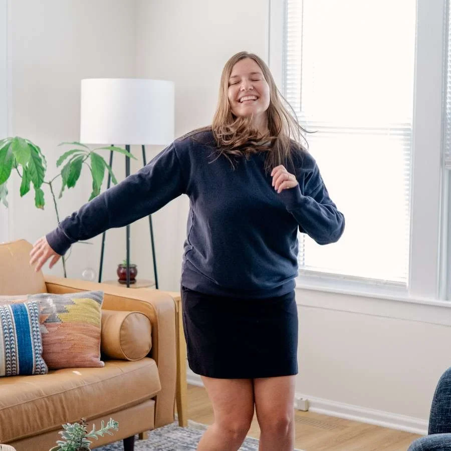 A woman dances joyfully inside a room with windows and a leather couch