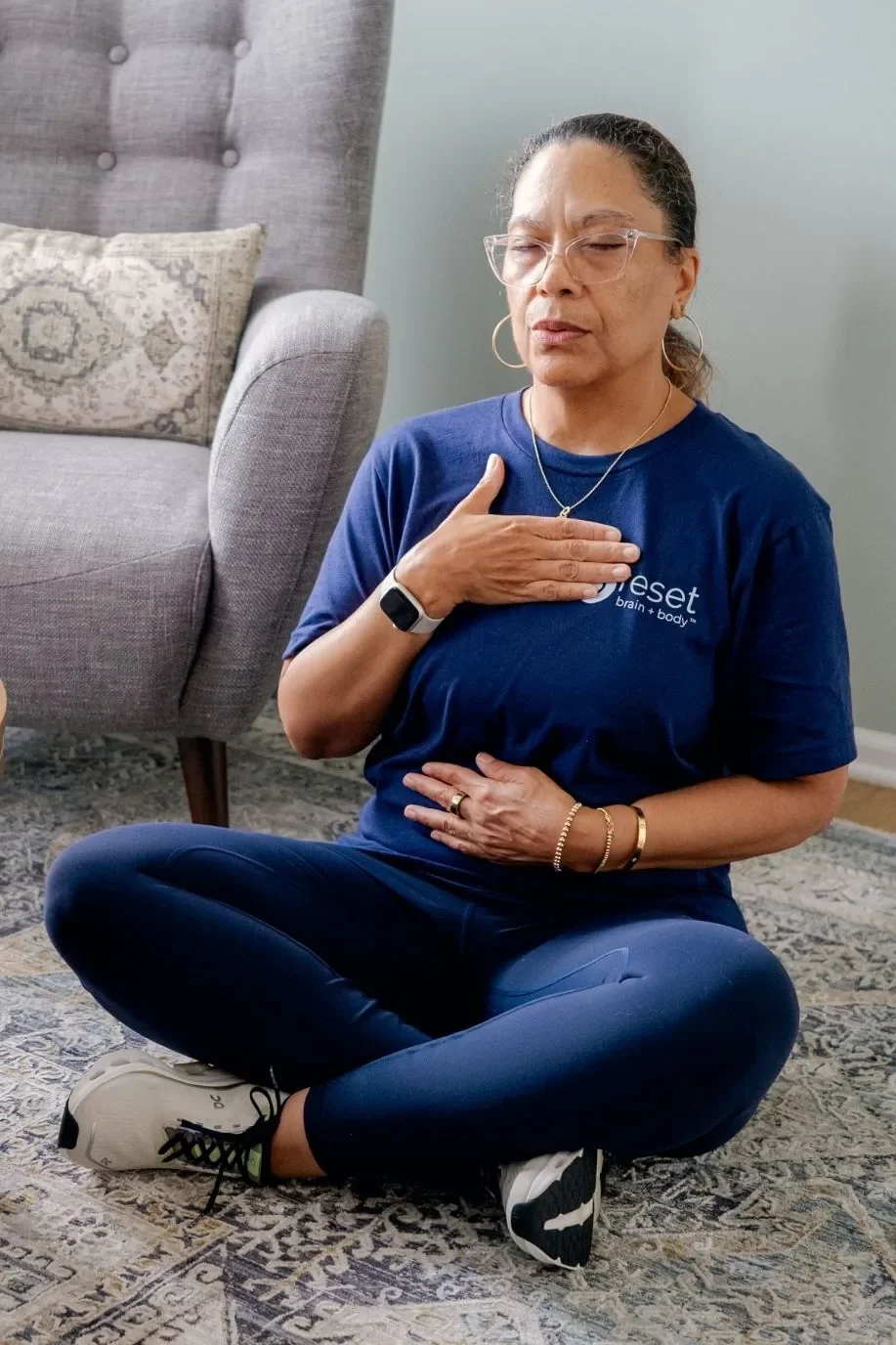 A woman practicing mindfulness or meditation indoors, sitting cross-legged with one hand on her chest and the other on her stomach, eyes closed, wearing casual athletic clothing.