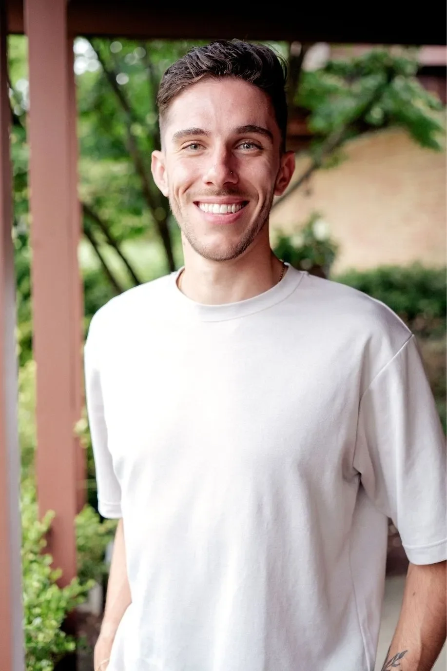 A man with a bright smile stands outdoors with greenery in the background.