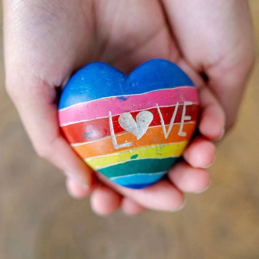 Two hands hold a heart-shaped stone painted in the colors of the rainbow with the word "Love" etched into the top