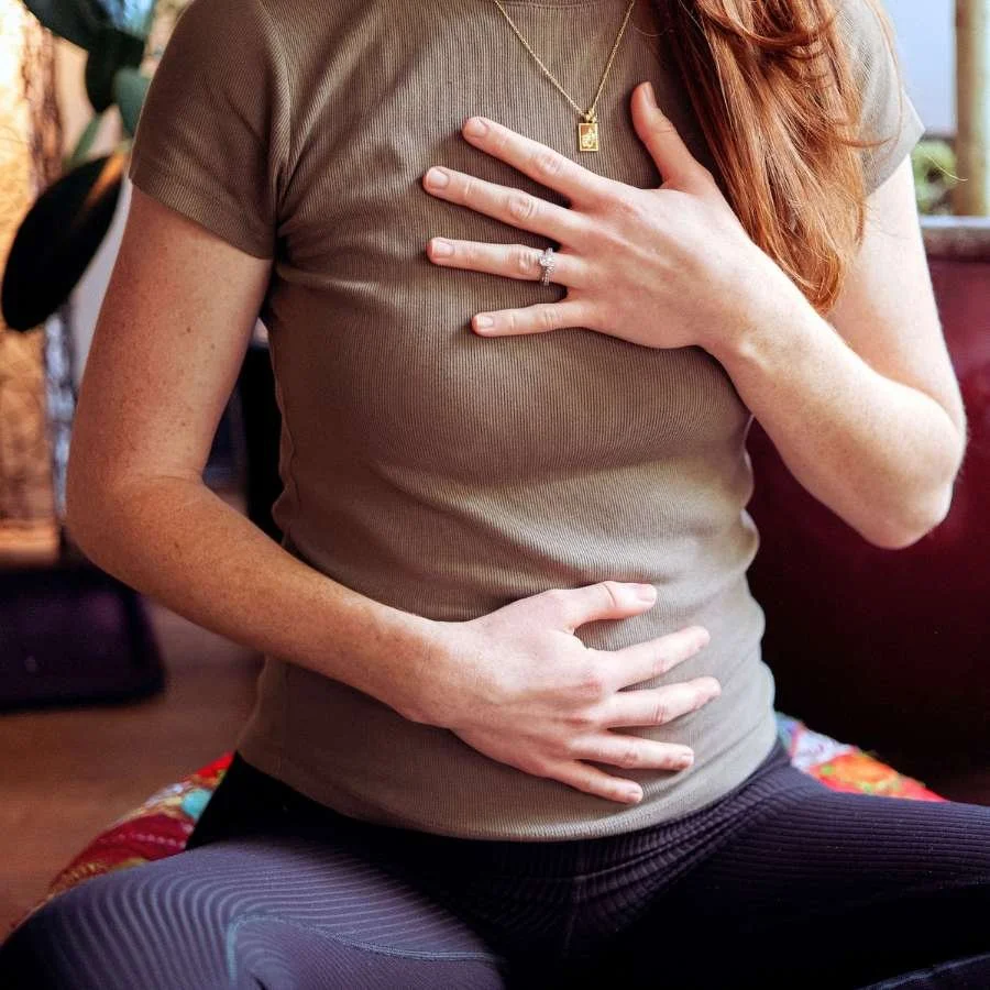 A red-haired woman sits in a cross legged position with hands over her heart and abdomen