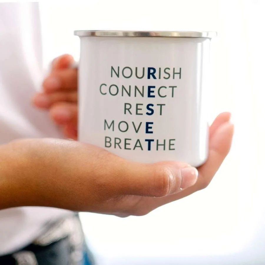 Someone holds a mug that lists the Reset Pillars of Wellness: Nourish, Connect, Rest, Move, Breathe.