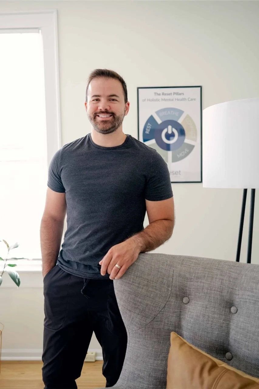 Blaise Fayolle, Reset therapist, smiles and stands in front of a gray chair. Behind him is a framed poster of the Reset Pillars of Foundational Wellness