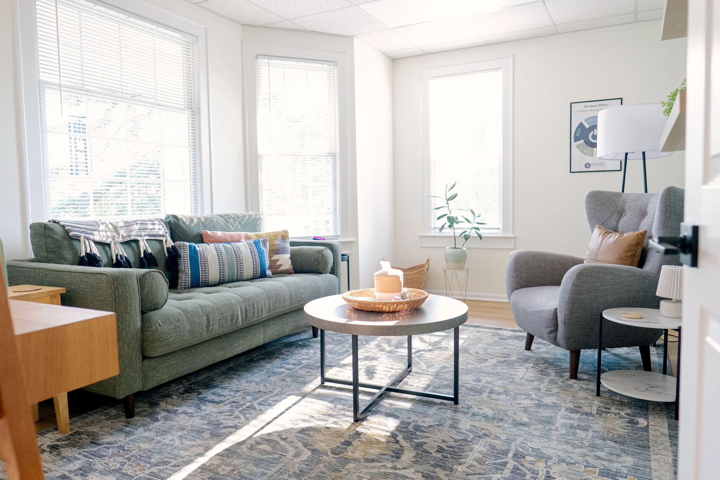 Cozy and inviting Reset office with a grey sofa, a grey armchair, a round coffee table, and large windows with white blinds, decorated with pillows, a plant, and a framed picture on the wall.