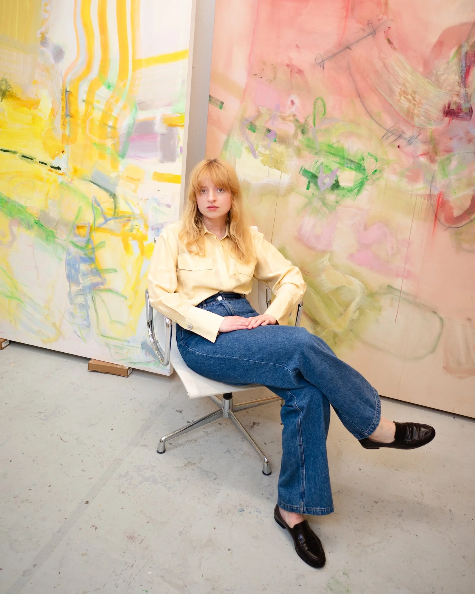 Artist Bunny Hennessey seated in a studio between two large abstract paintings in soft pink, yellow and green tones, surrounded by expressive layered brushstrokes and translucent marks.