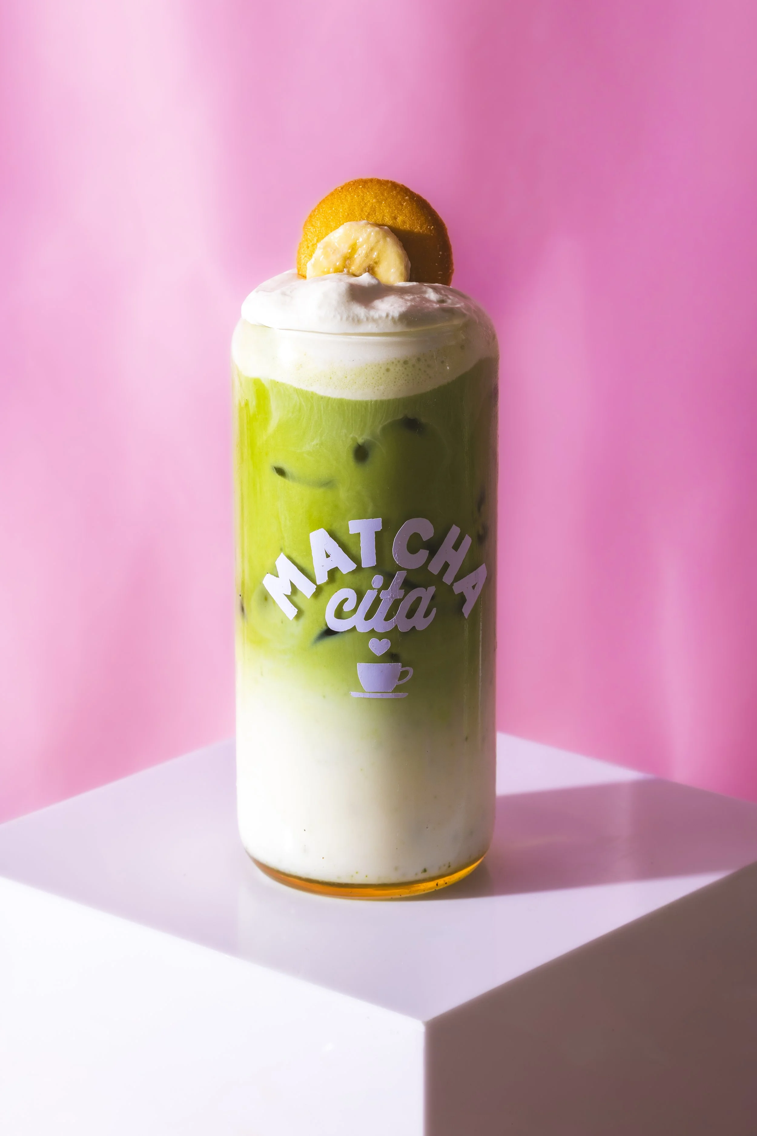 A layered matcha latte with whipped cream, a banana slice, and a cookie on top, in a clear glass with a "Matcha Cita" logo, set on a white surface with a pink background. Best banana matcha in Chicago, Fulton Market, Westloop