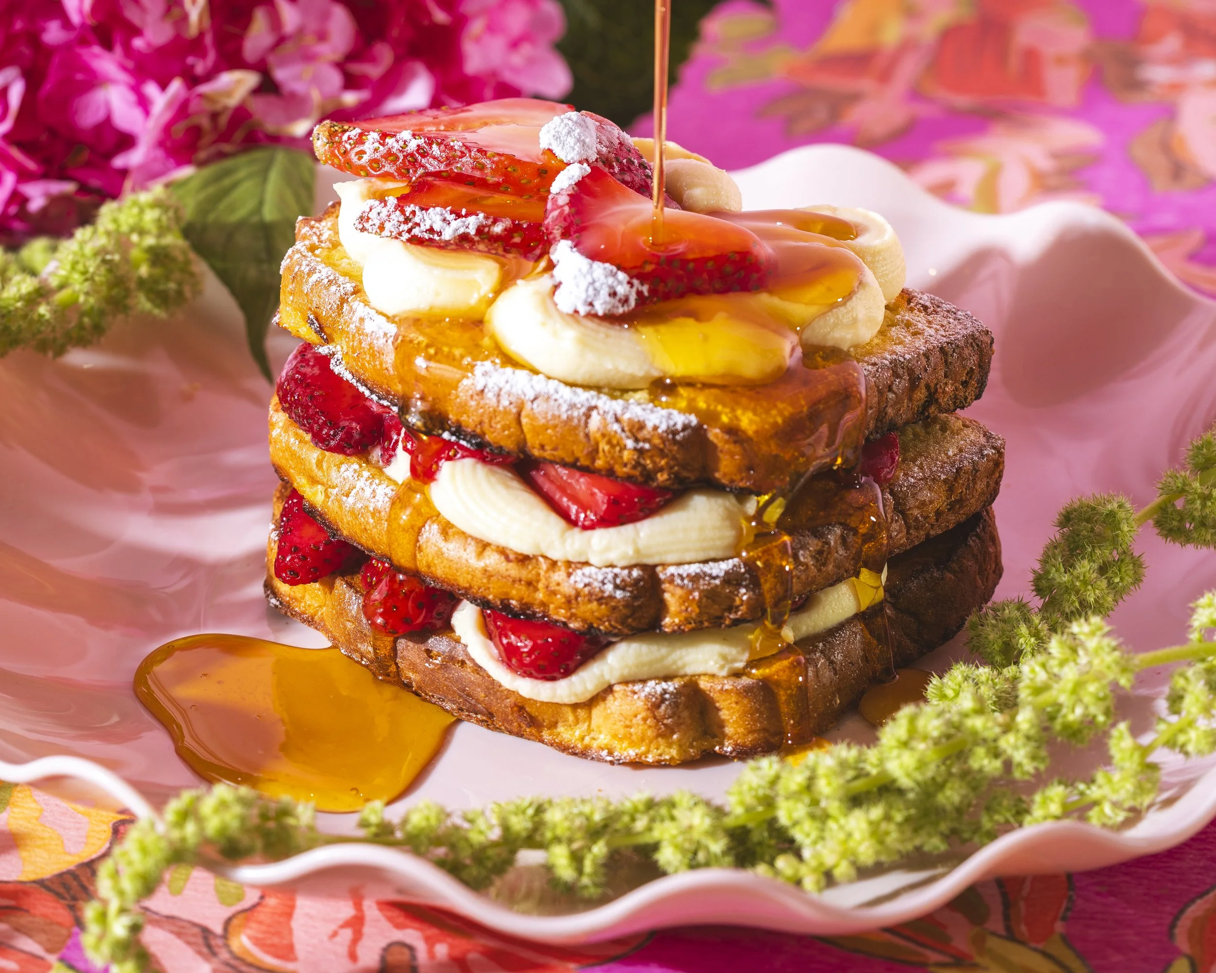 Layered French toast with strawberries, bananas, whipped cream, syrup, and powdered sugar on a pink plate with flowers in the background. Located at Matcha Cita, Fulton market cafe