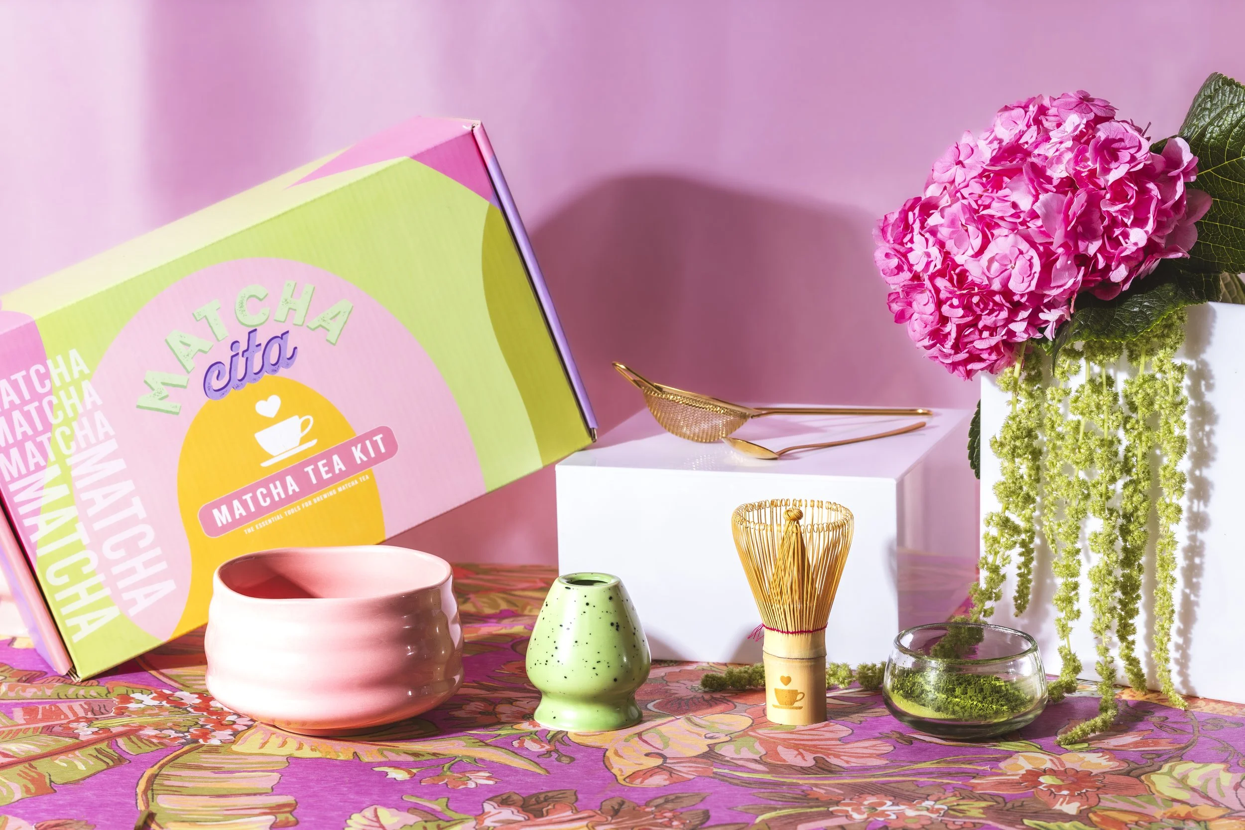 Colorful matcha tea kit with various tea accessories, a pink and green box labeled "Matcha Citta Matcha Tea Kit," a pink ceramic bowl, a small green ceramic container, and a bamboo whisk with a teacup icon, all arranged on a floral tablecloth with pi