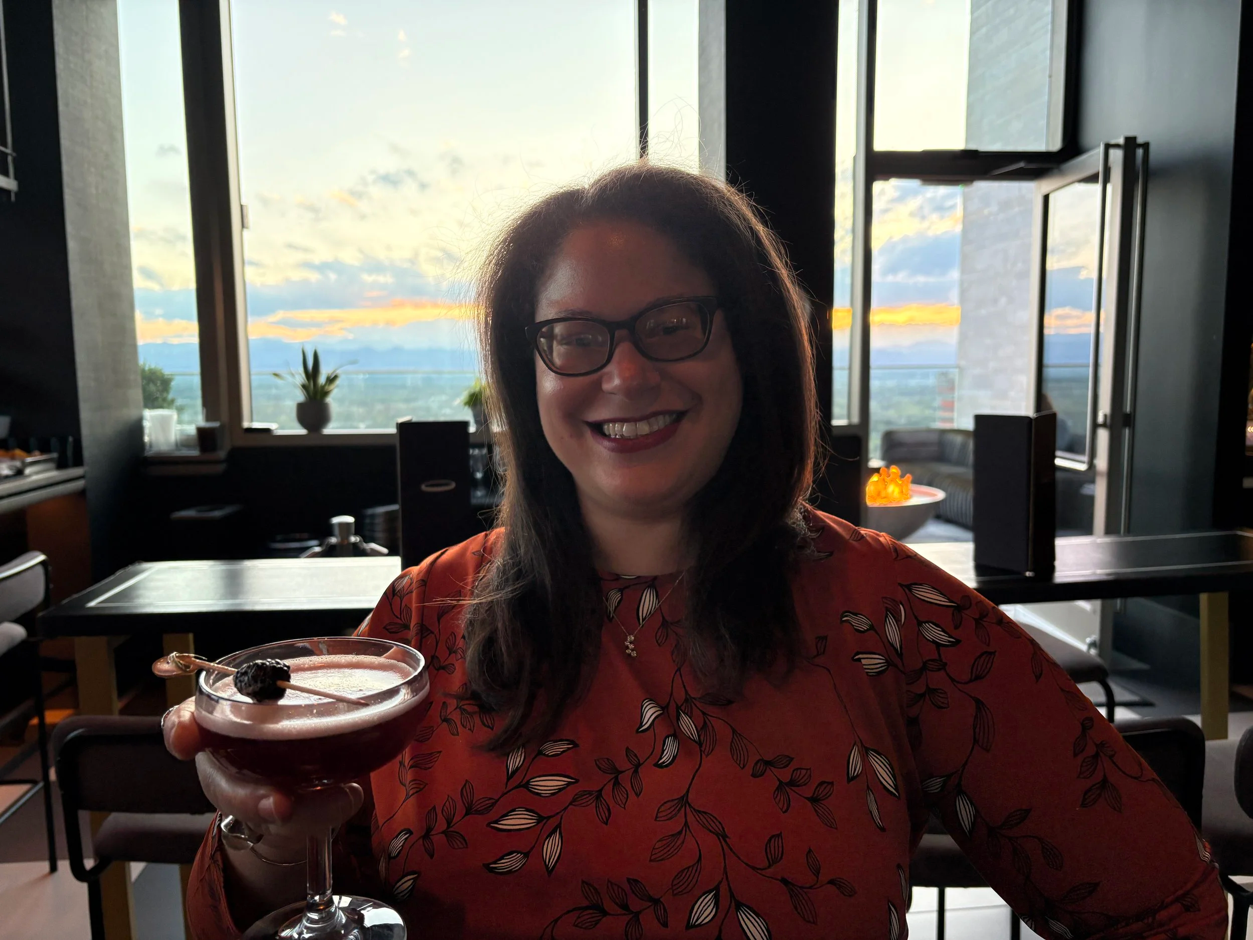 A woman with dark hair, glasses, and a red patterned blouse smiling while holding a cocktail glass with a dark beverage and garnished with a cherry, sitting in a restaurant with large windows and a view of the sky during sunset.