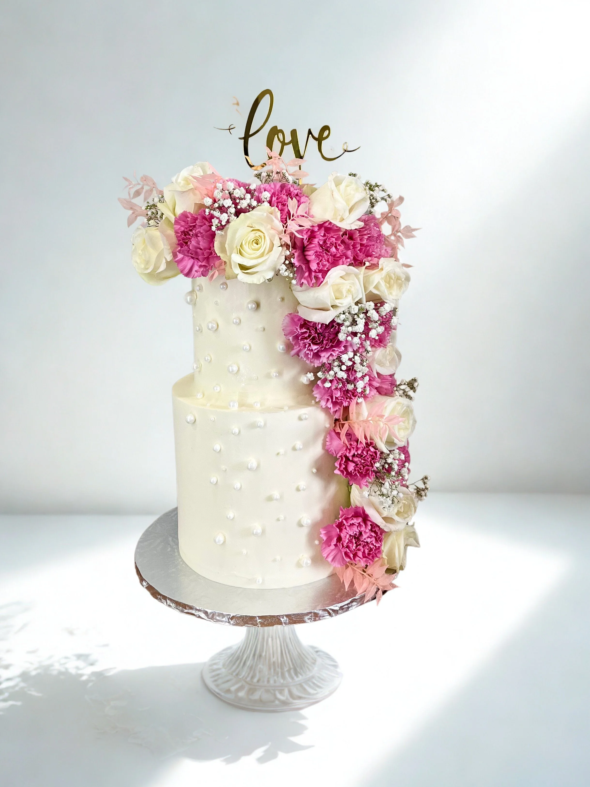 Elegant two-tier wedding cake decorated with white and pink flowers, pearl accents, and a gold "love" topper, displayed on a white cake stand.