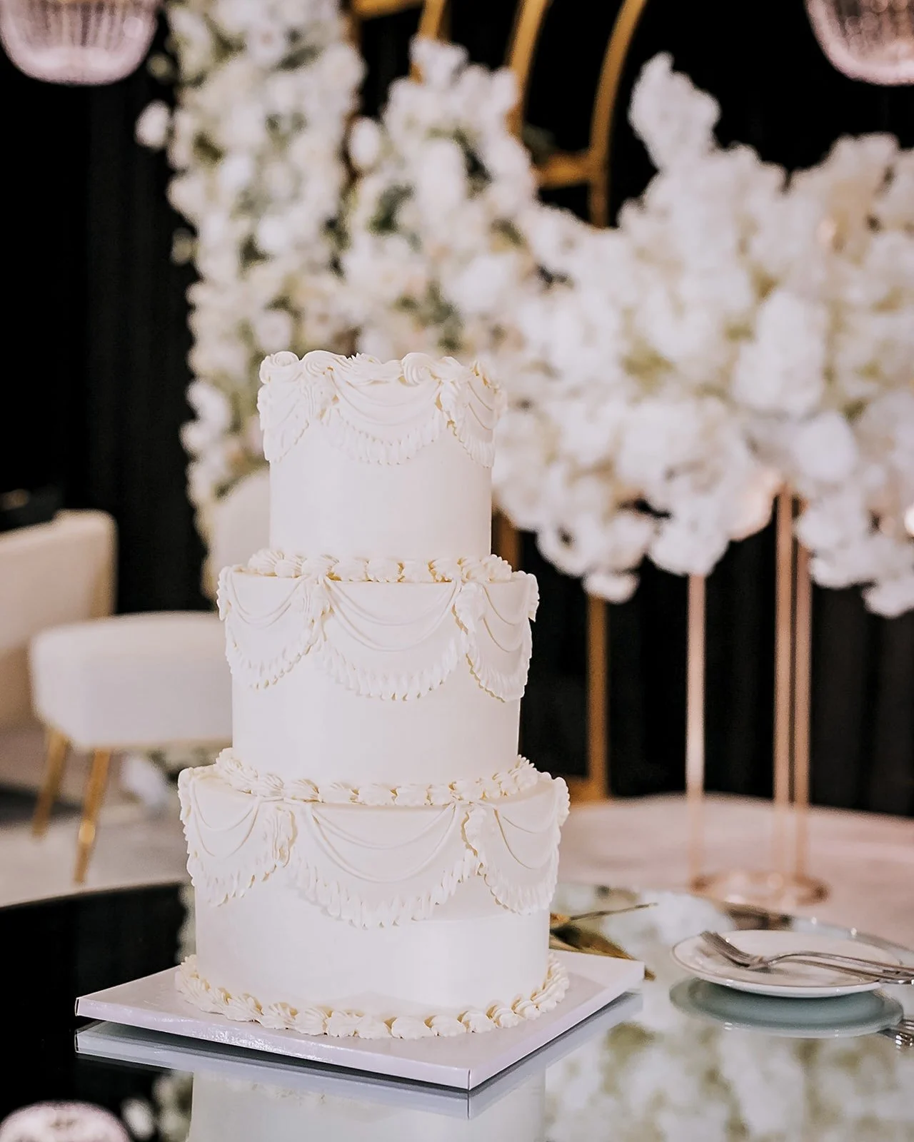 Three-tier white wedding cake with elegant piping and decorative drapes, set on a reflective surface with plates and utensils nearby, against a backdrop of large white floral arrangements.