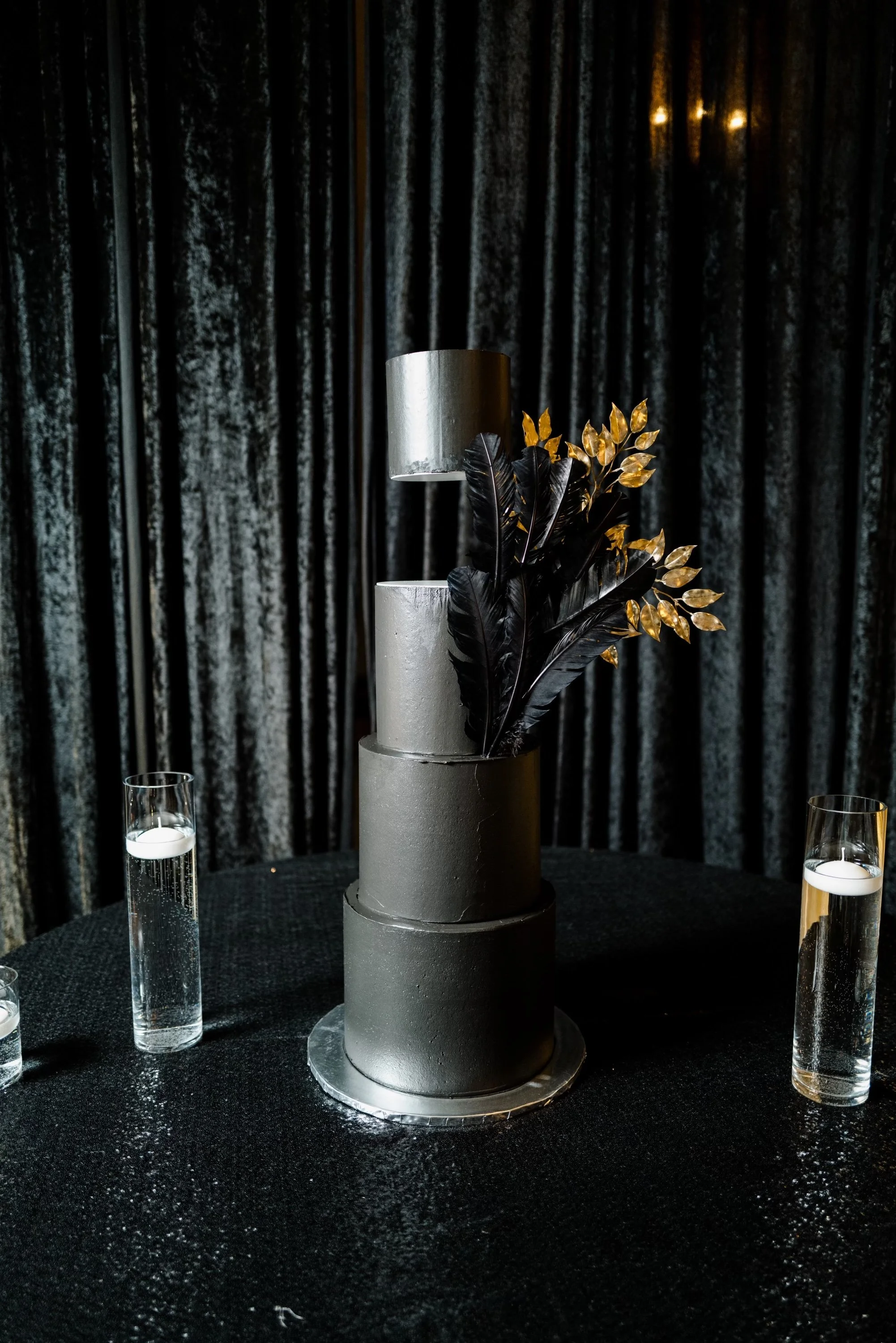 A four-tier black wedding cake with gold and black decorative foliage, placed on a round black table with three glasses of champagne, against black velvet curtains.