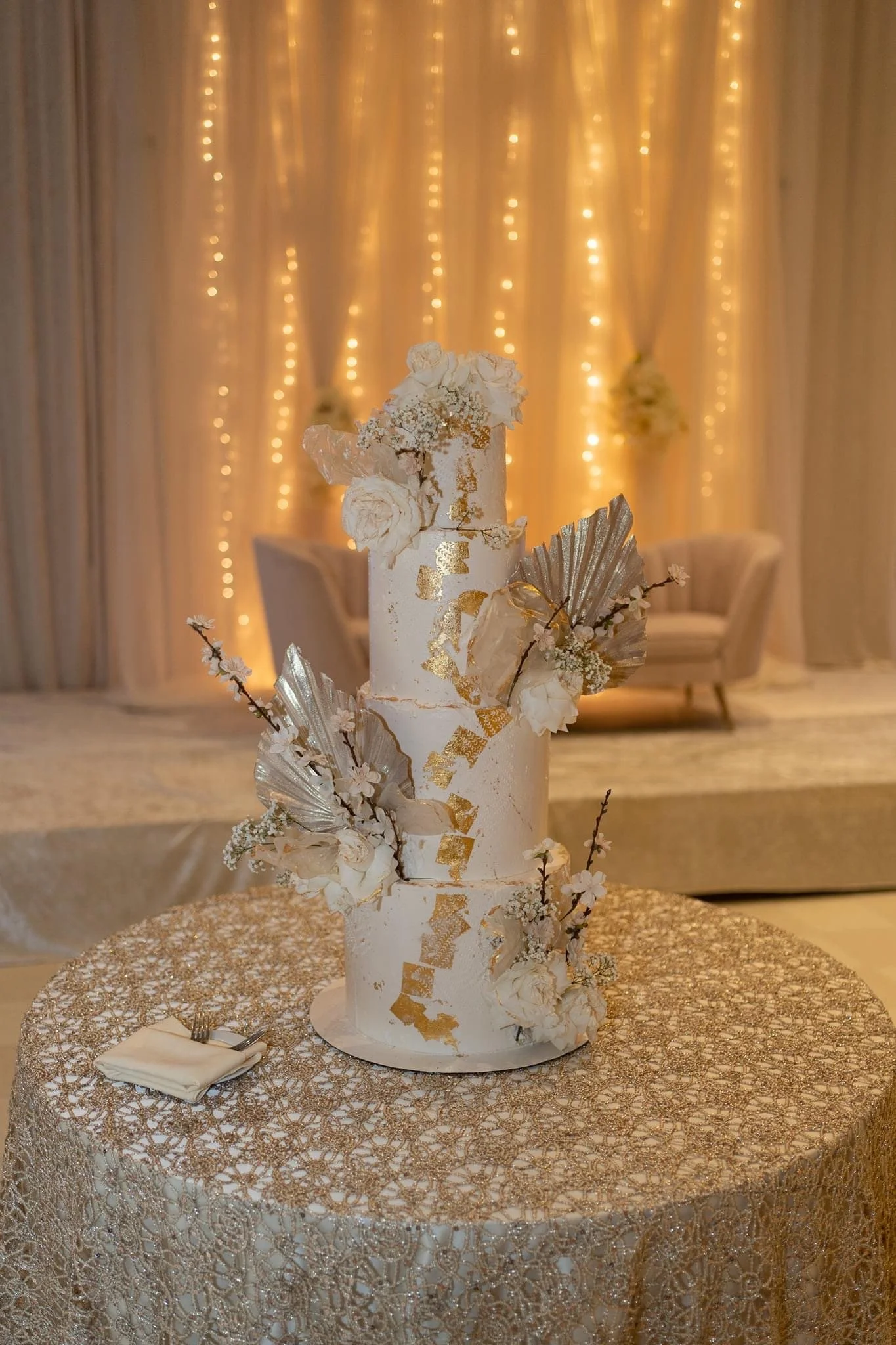 A three-tier white wedding cake decorated with gold accents, white flowers, and metallic leaves, placed on a lace tablecloth at a decorated reception table.