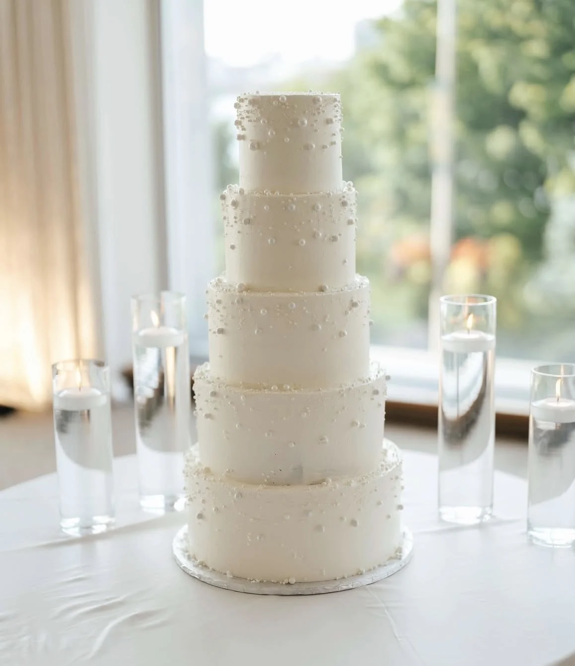 A tall, multi-tiered white wedding cake with pearl-like decorations on each tier, placed on a white table, with five lit candles in glass holders surrounding it, and a large window with greenery outside in the background.