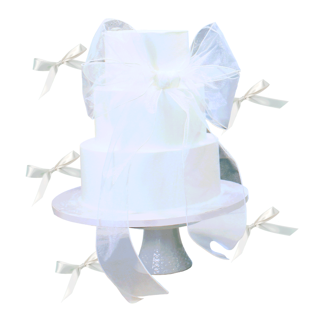White, two-tiered cake on a white cake stand decorated with white ribbons and bows, set against a plain background.