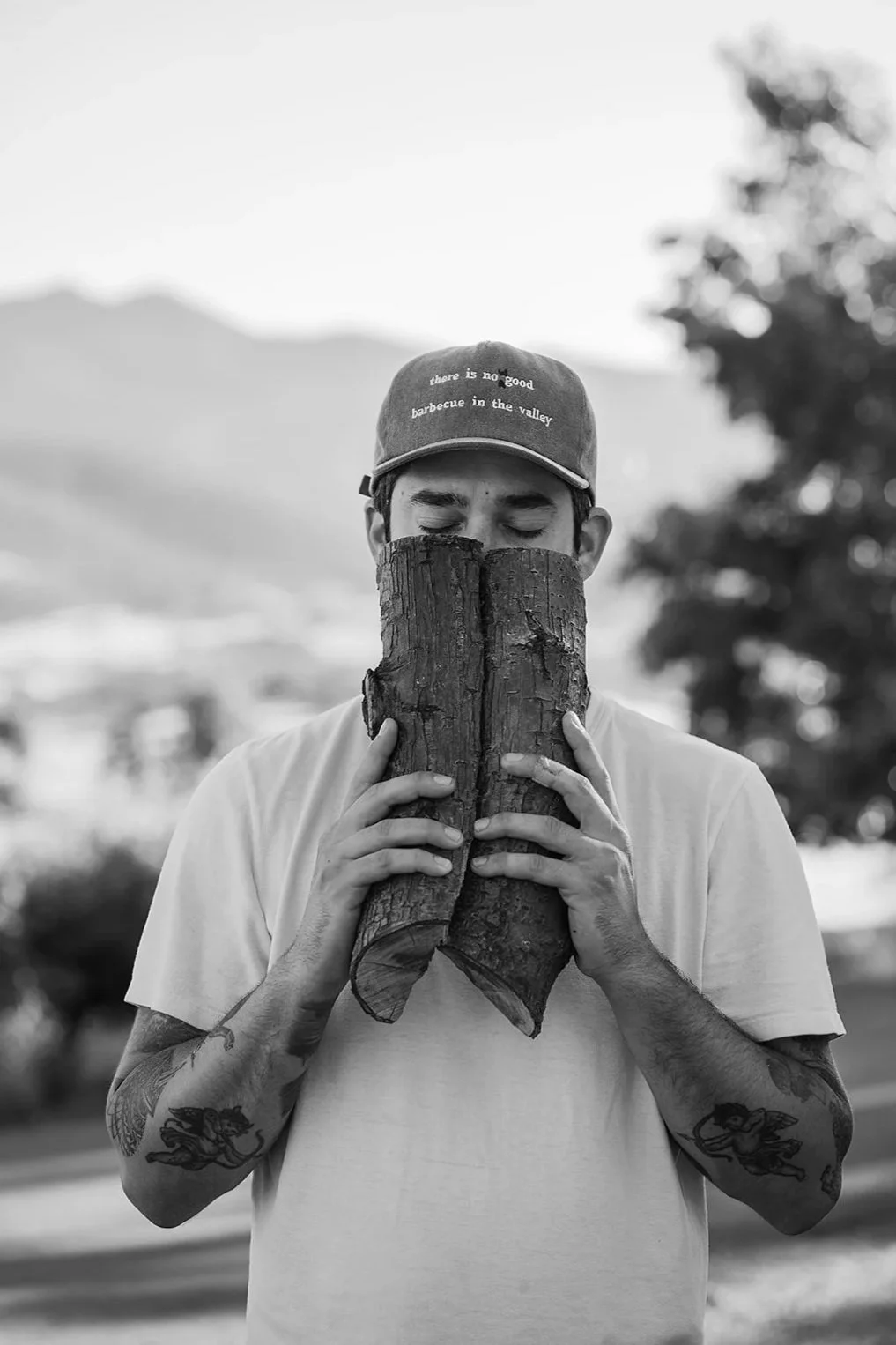 A person holding two pieces of wood in front of their face with eyes closed, wearing a cap with text, outdoors with trees and mountains in the background.