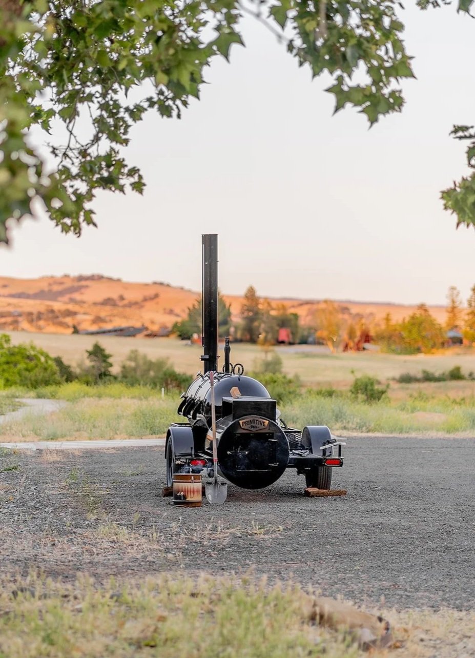 A black smoker grill on a trailer parked on a gravel surface, with a scenic landscape of hills, trees, and a clear sky in the background.
