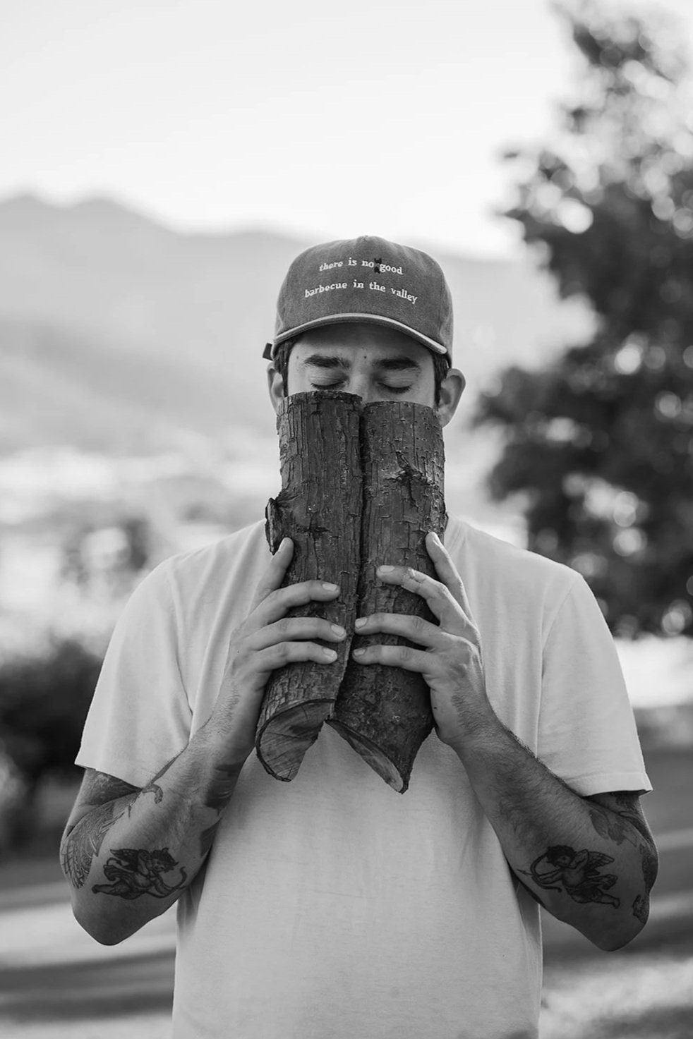 A person holding two pieces of wood in front of their face with eyes closed, wearing a cap with text, outdoors with trees and mountains in the background.