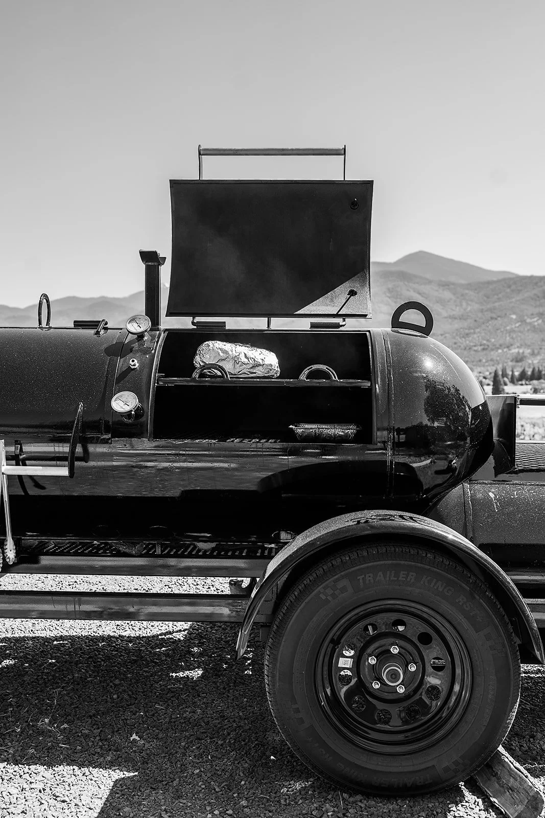 A black barbecue grill on a trailer with a mountain background, featuring a foil-wrapped item inside, in black and white.