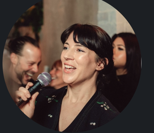 A woman holding a microphone and smiling during an event with people in the background.