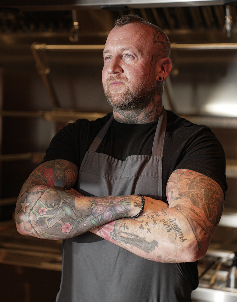 Tattooed man with short hair and beard wearing a black shirt and gray apron, standing with arms crossed in a woodworking shop.
