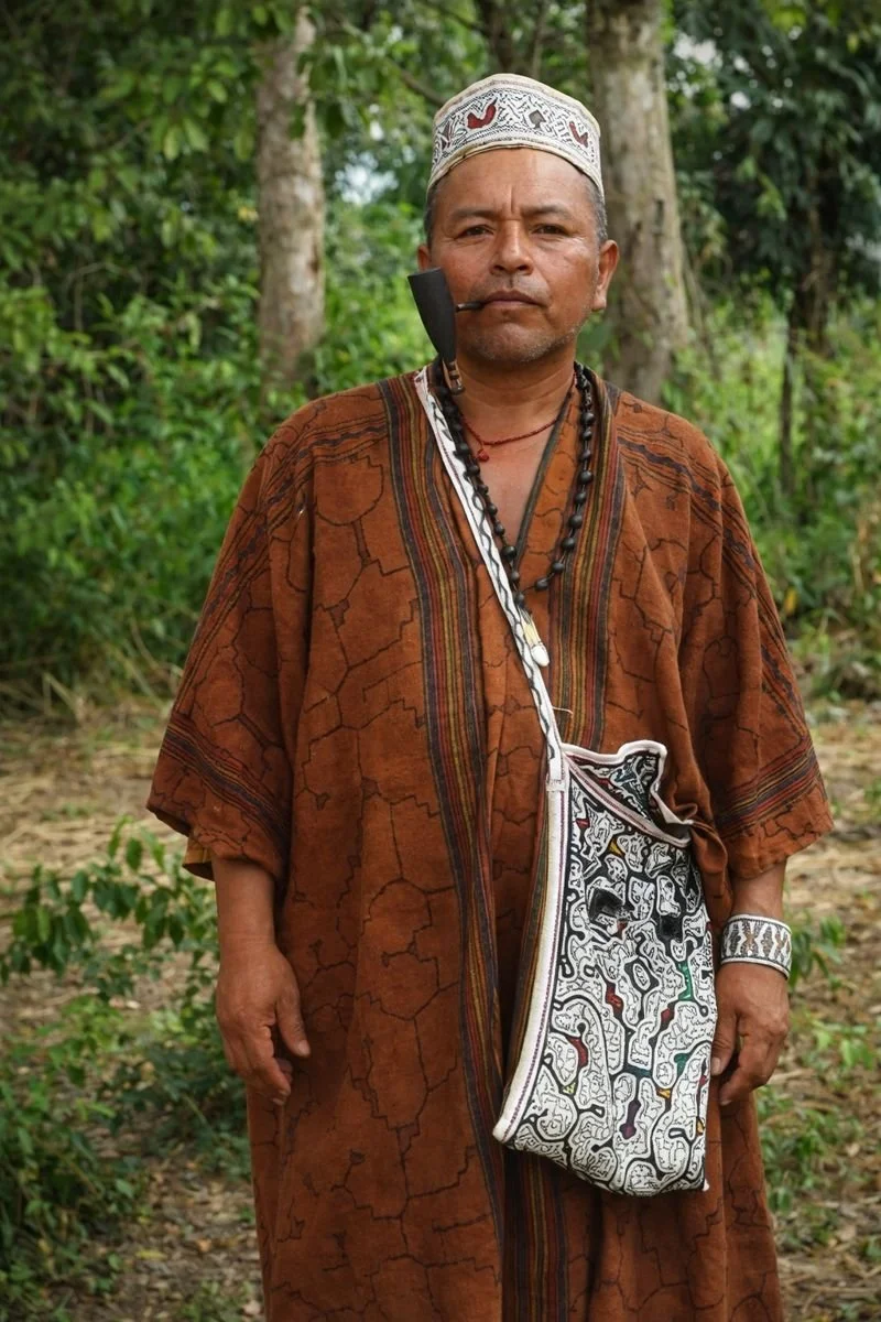 A man standing outdoors in a forested area, wearing traditional tribal attire with a pipe in his mouth, a beaded necklace, a patterned bag, and a decorated hat.