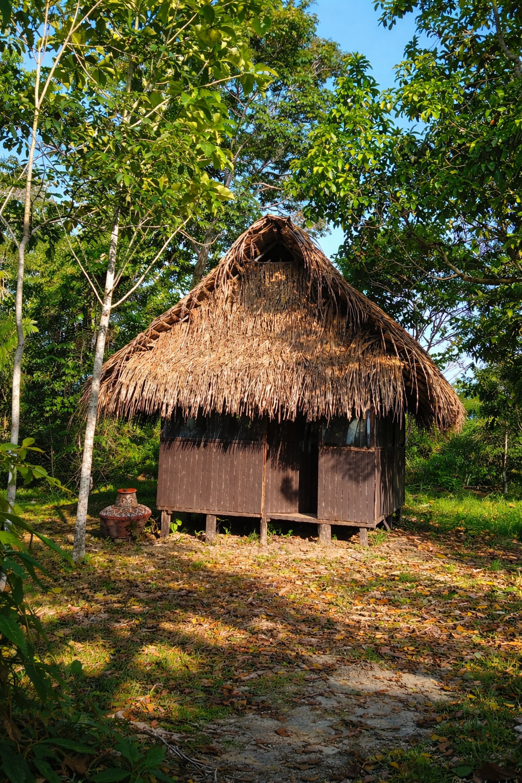 A small traditional hut with a thatched roof made of palm leaves, surrounded by lush green trees, with a decorative clay pot to the left of the hut and a ground covered in fallen leaves.