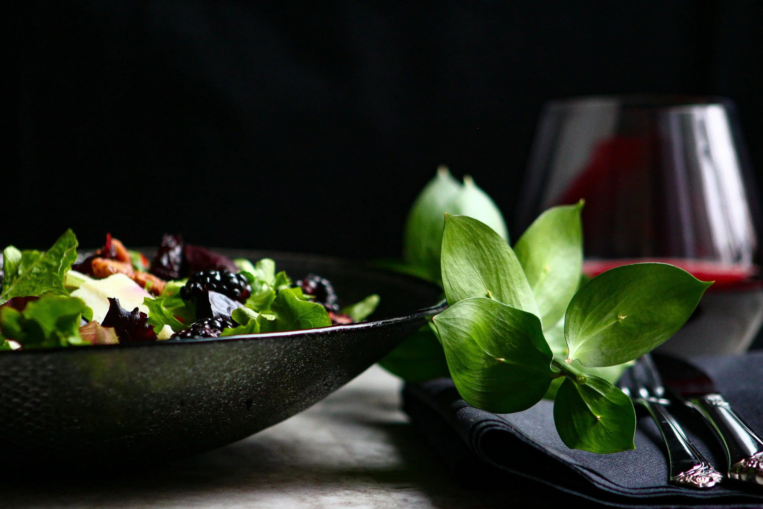 Close-up of a salad in a black bowl with greens, blackberries, and other berries, with green leaves and a glass of red wine in the background on a dark setting.