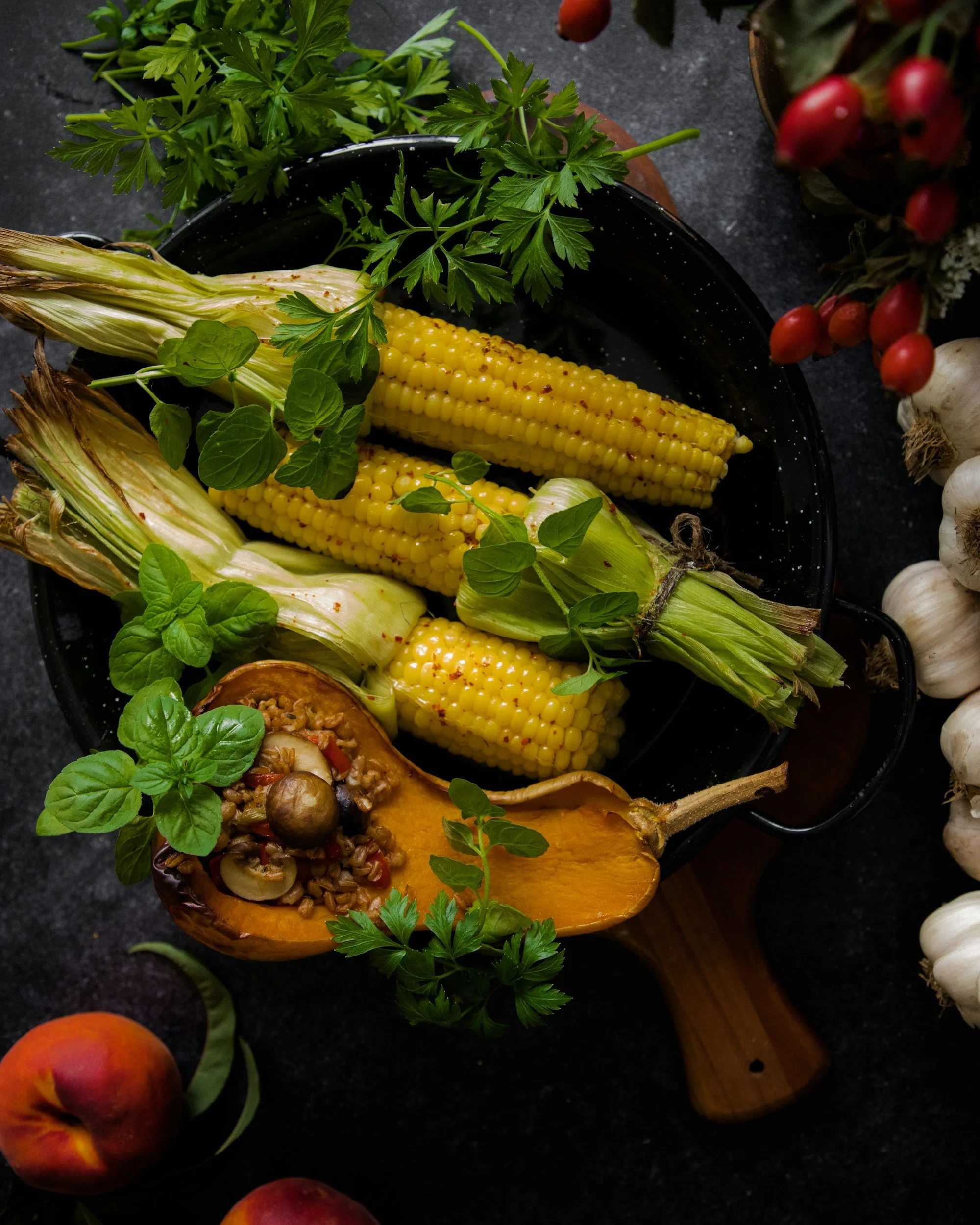 Corn on the cob, fresh herbs, a halved pumpkin, garlic, and apples arranged on a dark surface.
