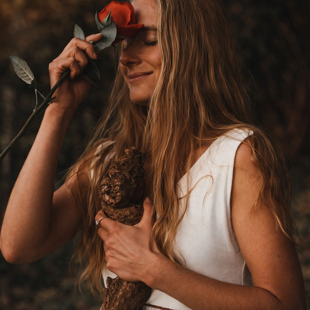 A woman with long, wavy red hair holding a large mushroom close to her chest with her left hand, and a red rose in her right hand pressing against her forehead, standing outdoors with a blurry natural background.