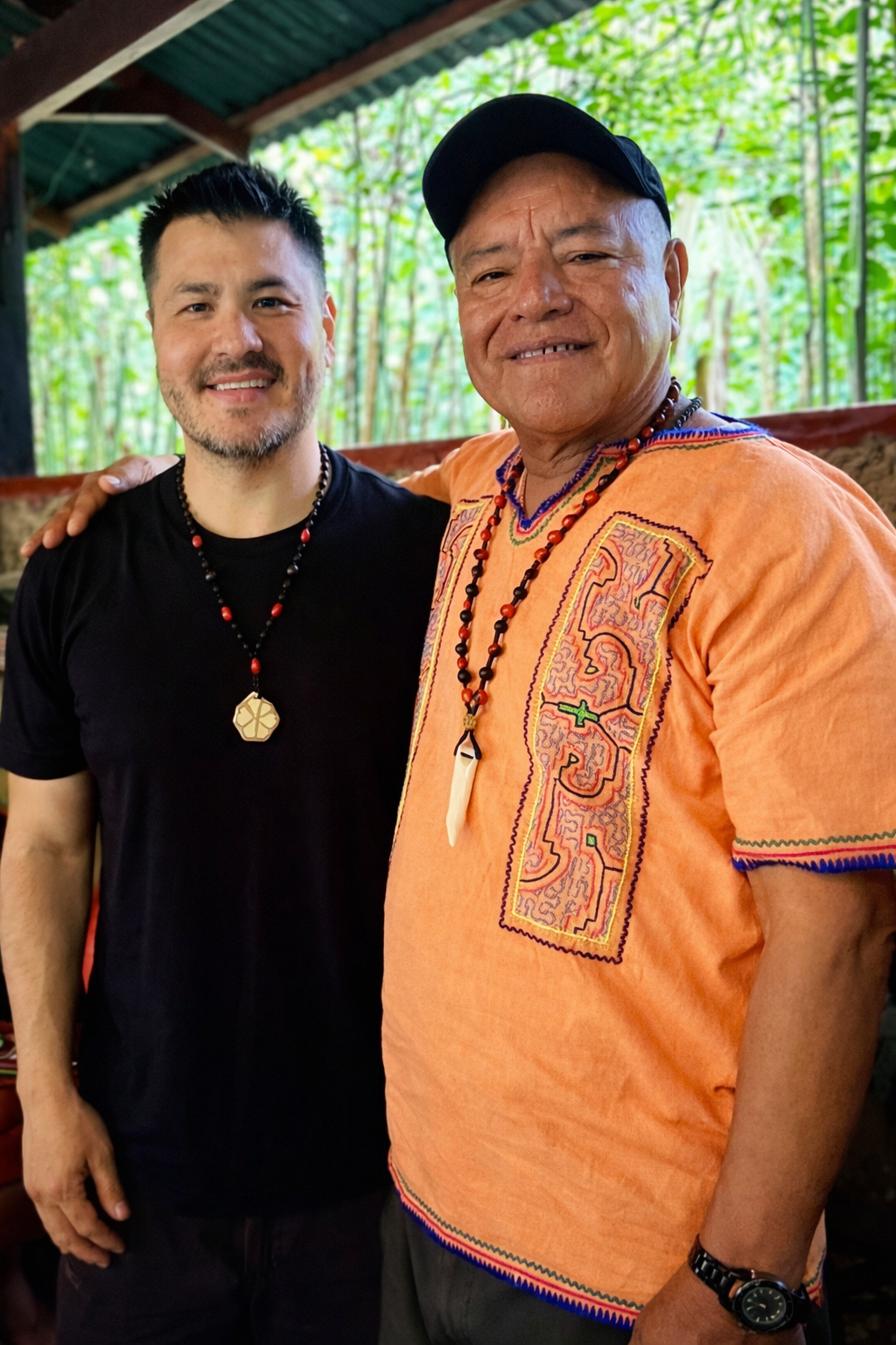 Two men standing side by side outdoors, smiling. One is wearing a black shirt and necklace, the other is wearing an orange embroidered shirt, a black cap, and beaded necklaces, with tropical greenery in the background.