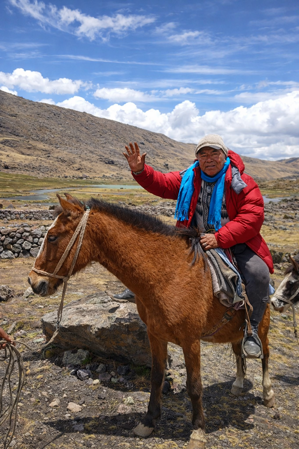 Man in a red jacket and blue scarf riding a brown horse in a mountainous landscape with clouds in the sky.