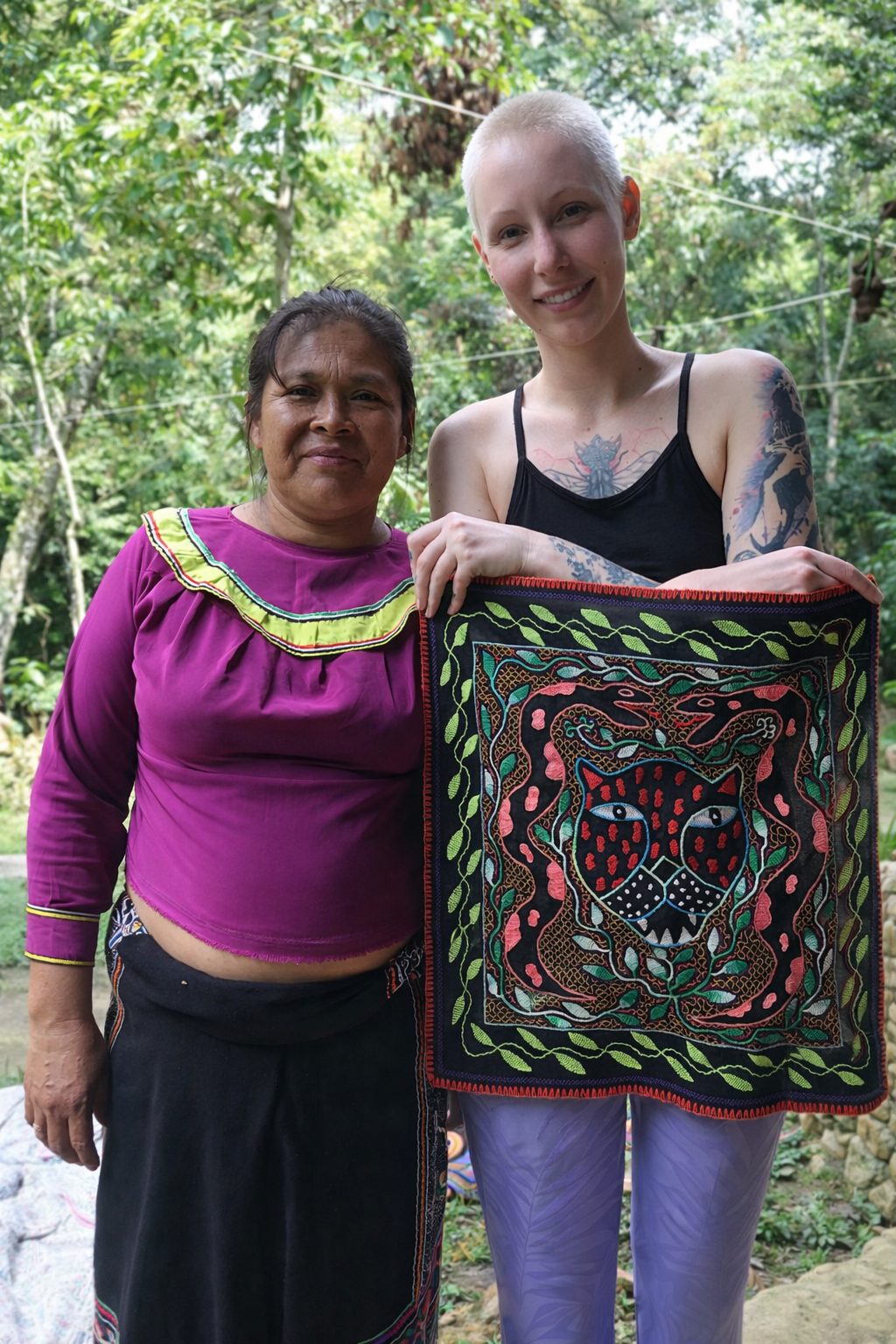 Two women standing outdoors in front of green trees, holding a colorful embroidered textile with a black panther and animal motifs.