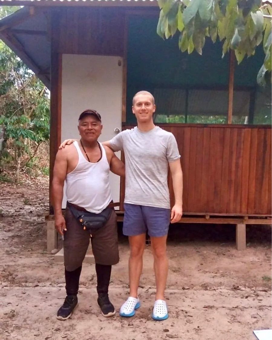 Two men standing side by side outdoors in front of a small wooden structure, smiling at the camera. One man is wearing a white tank top, dark shorts, and a black cap, while the other is wearing a light gray t-shirt, blue shorts, and white Crocs.